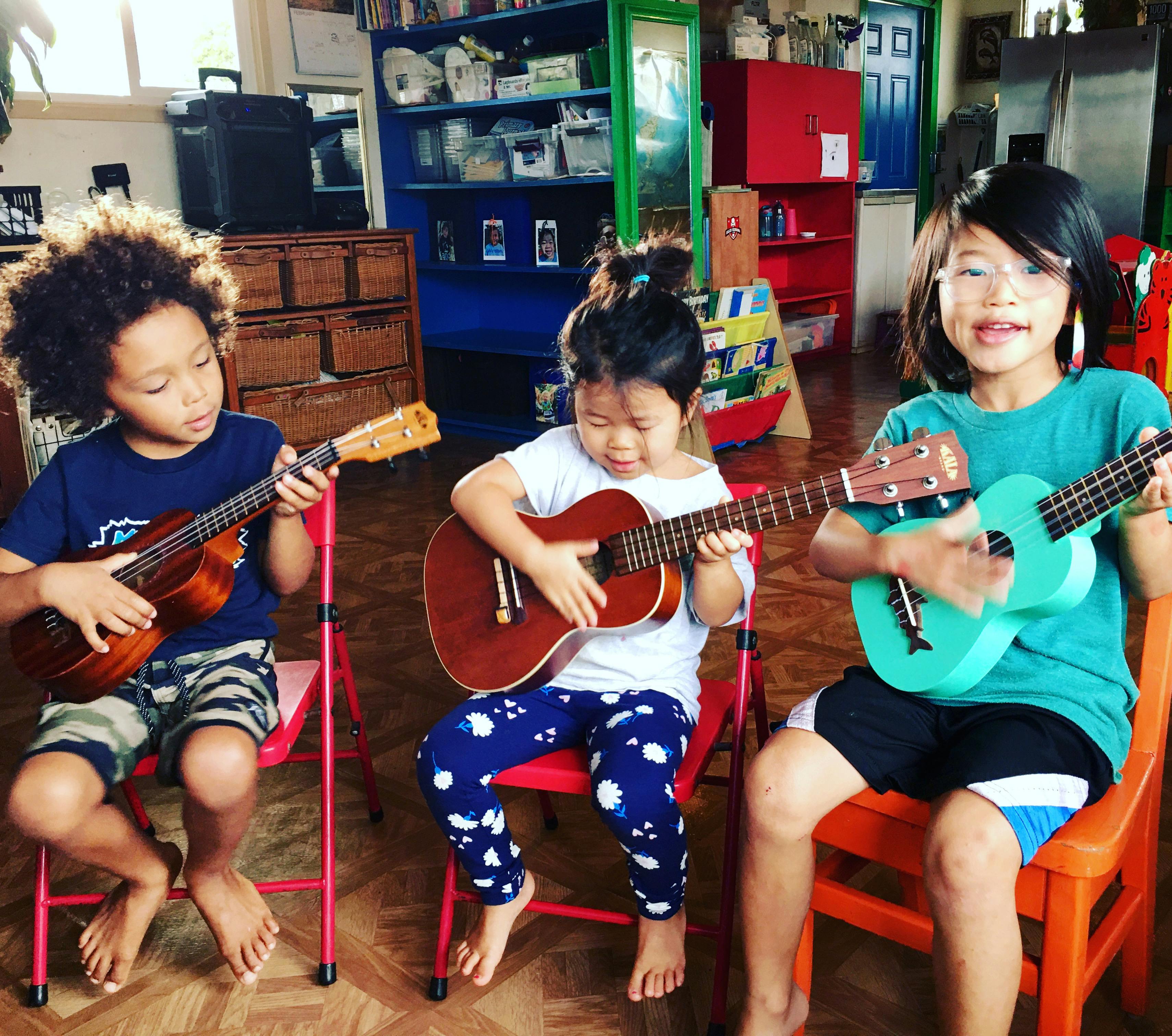 Three children sitting on chairs playing ukuleles