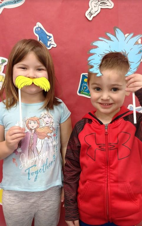 Two children holding up props to their faces paper cutouts of a yellow mustache and blue hair