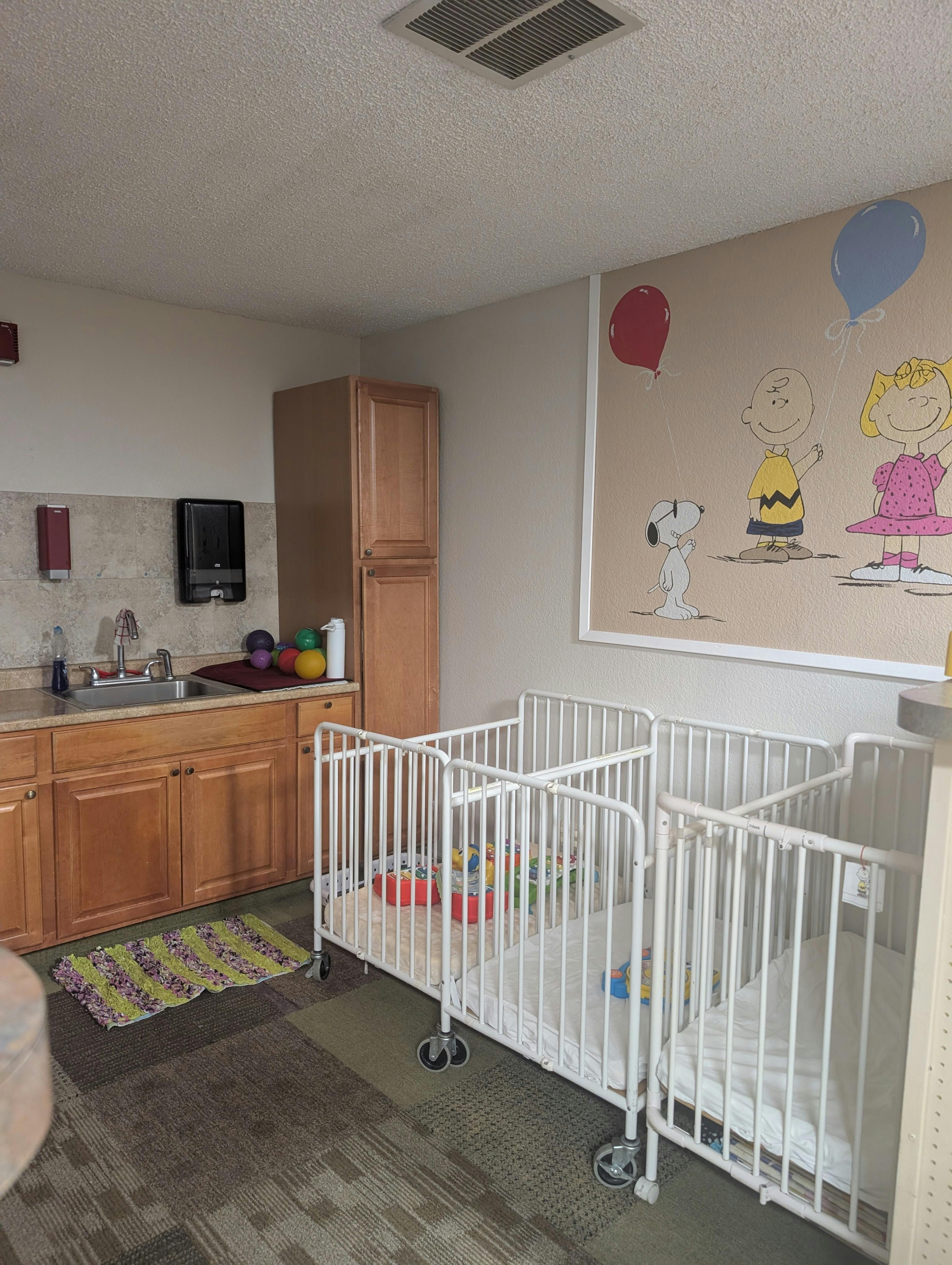 Cribs in a daycare room next to a kitchen counter and cartoon wall mural