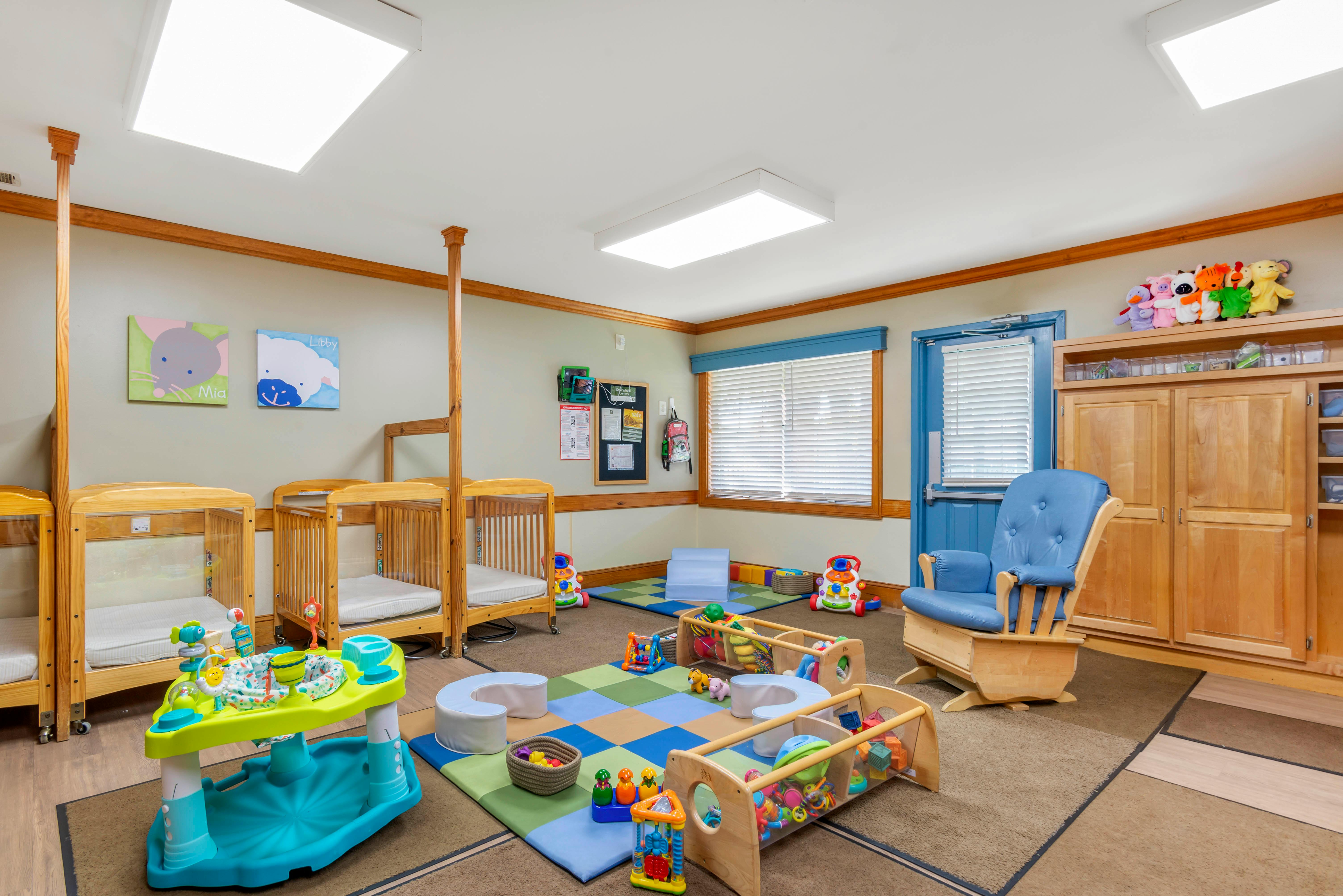 A brightly lit nursery room with multiple cribs, a rocking chair, and play mats