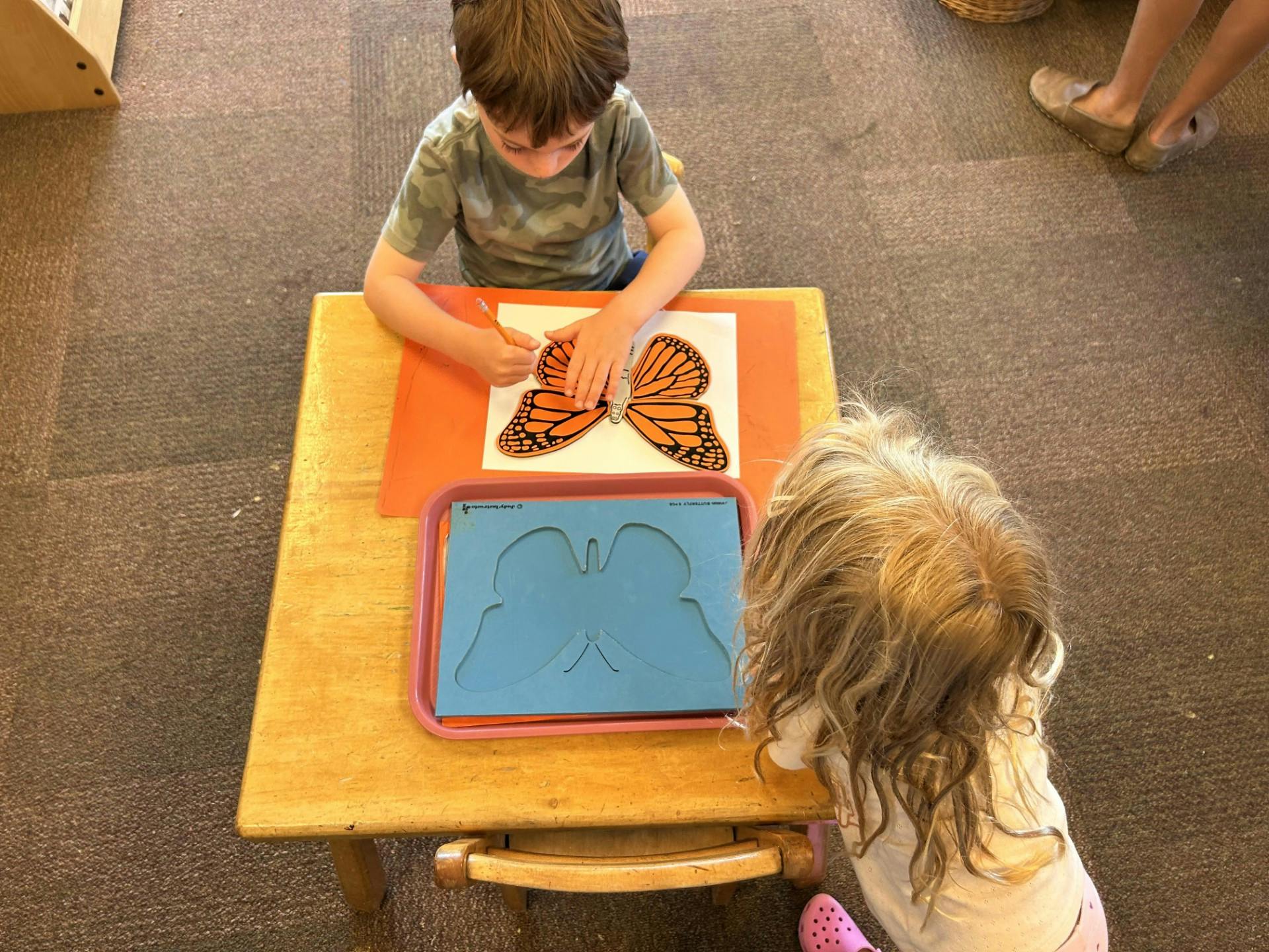 Child tracing a butterfly shape with another child observing