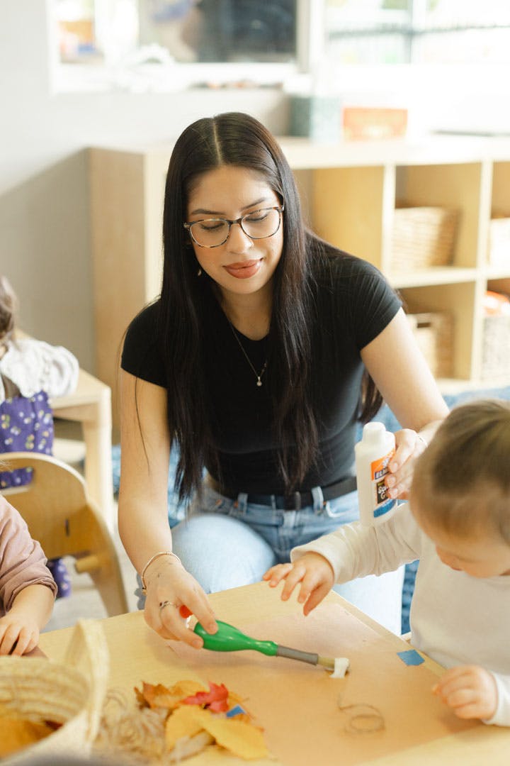 Person helping a child glue leaves on paper at a table