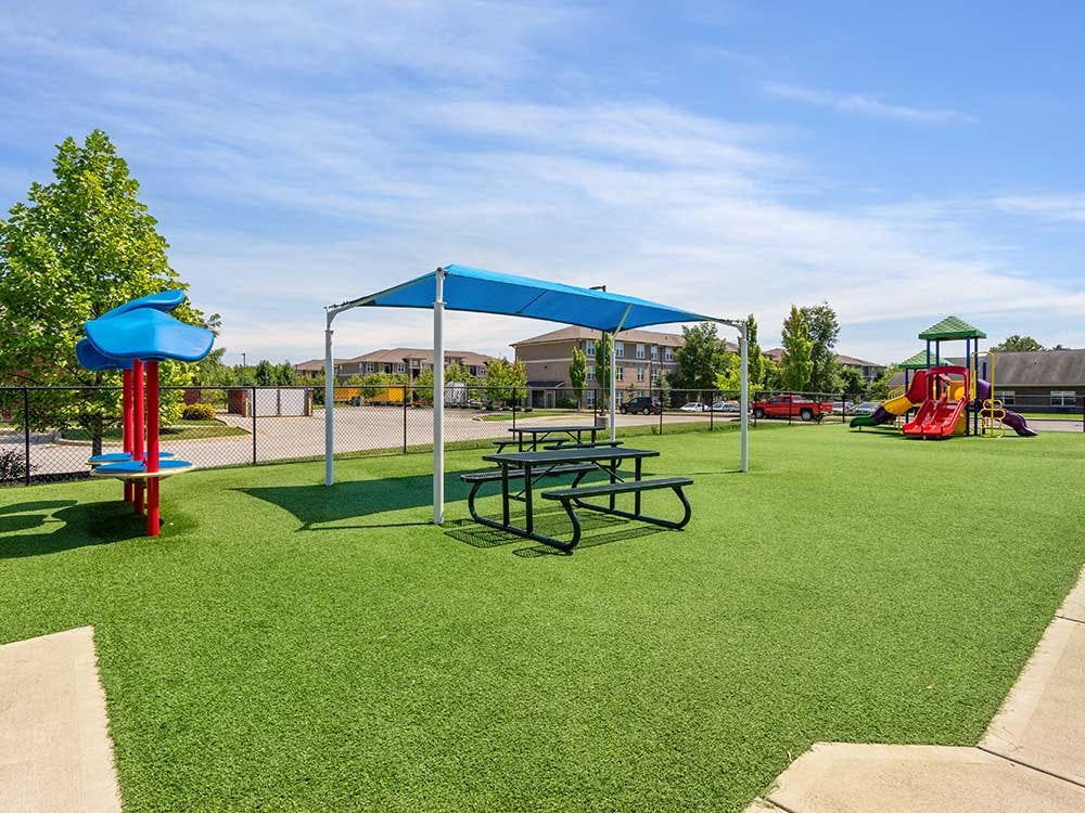 Playground area with green turf, a blue shade, picnic tables, and play structures