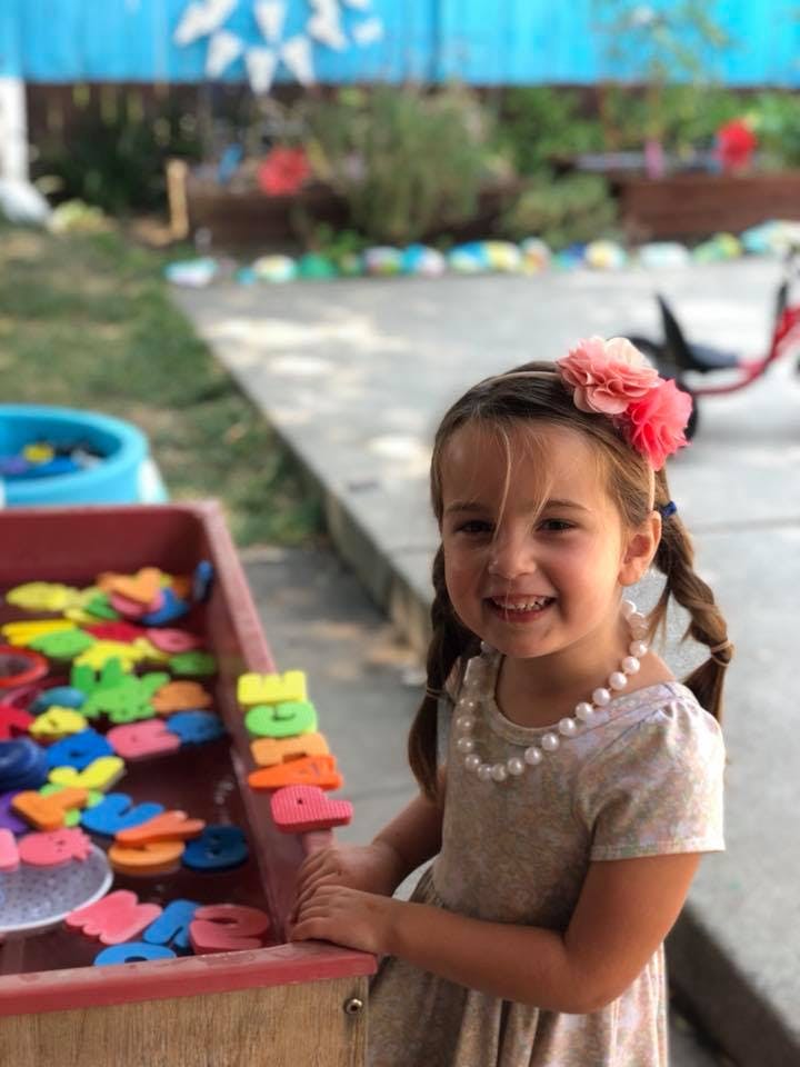 Child with pigtails and a flower headband standing next to a bin of colorful letters