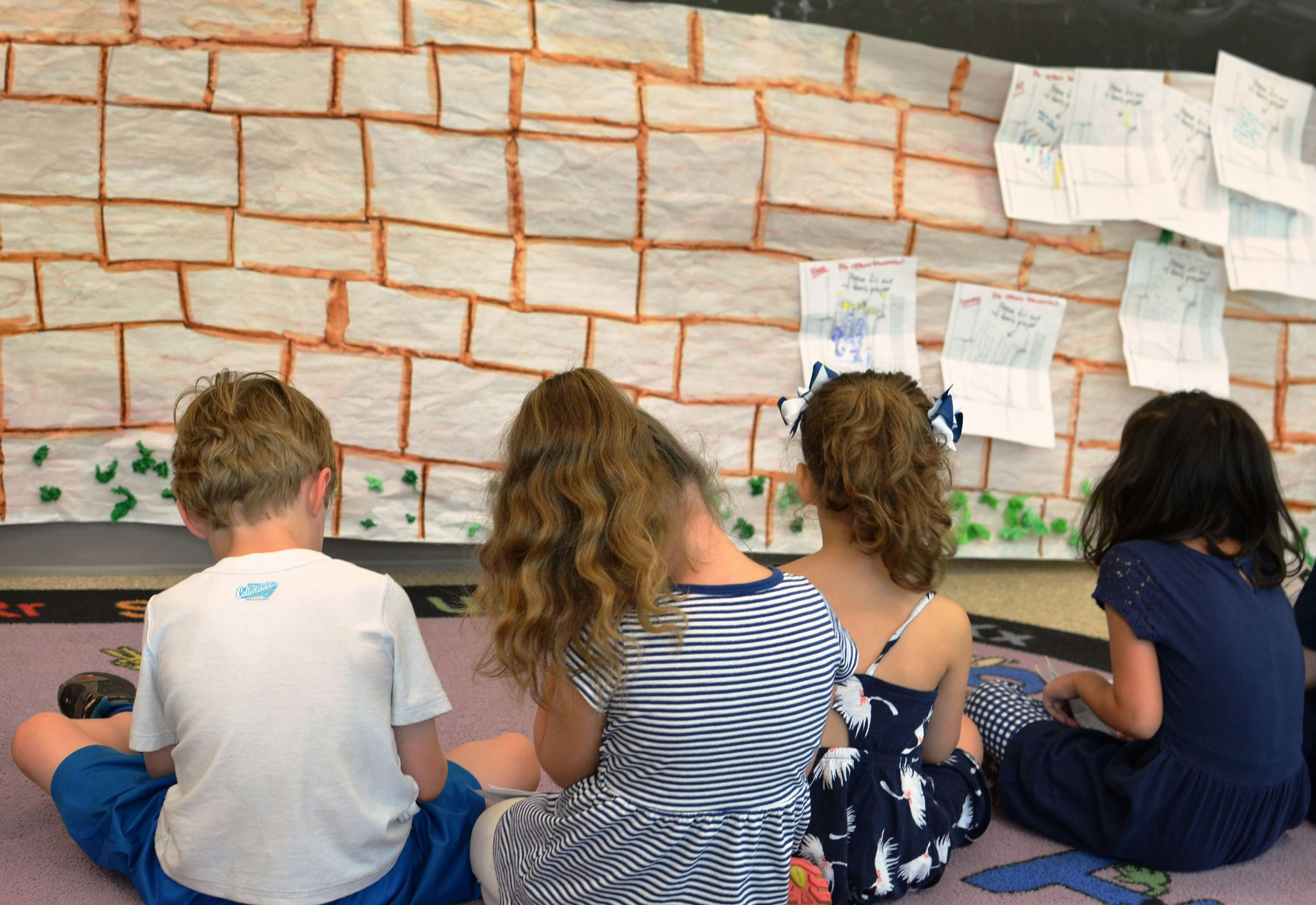 Children sitting on the floor facing a wall with paper brick art