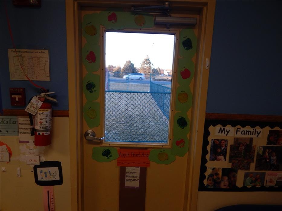 Classroom door decorated with paper apples, looking out through window to fence and road
