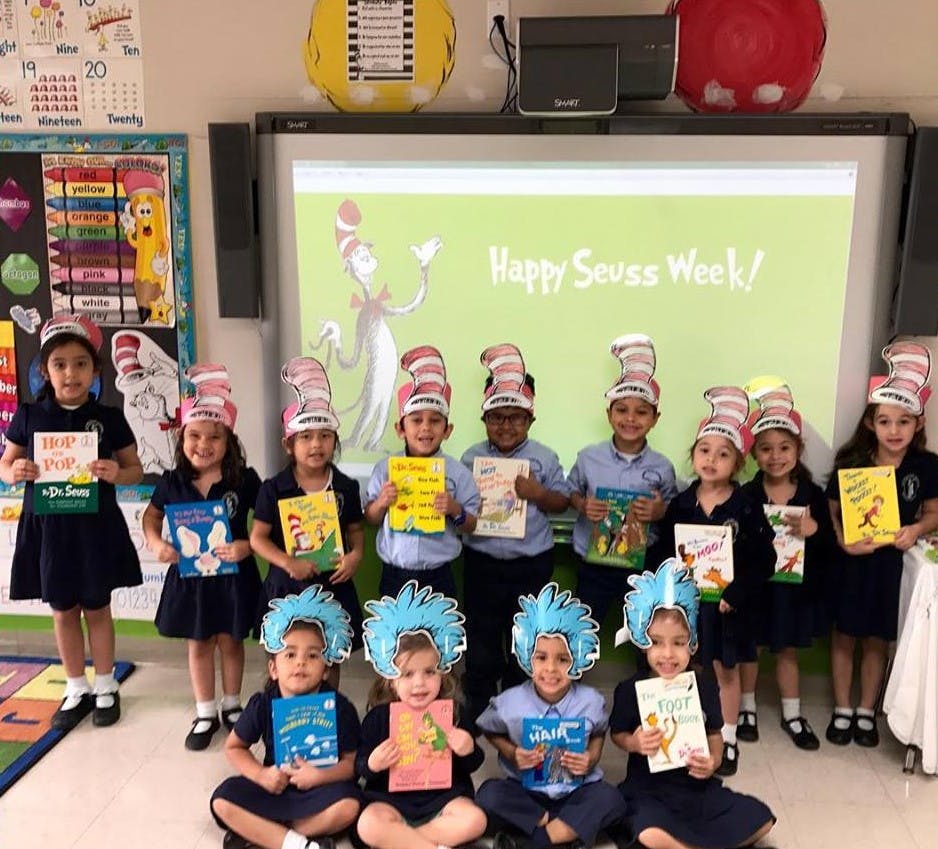 Children in a classroom wearing hats and holding books, with "Happy Seuss Week!" on a screen