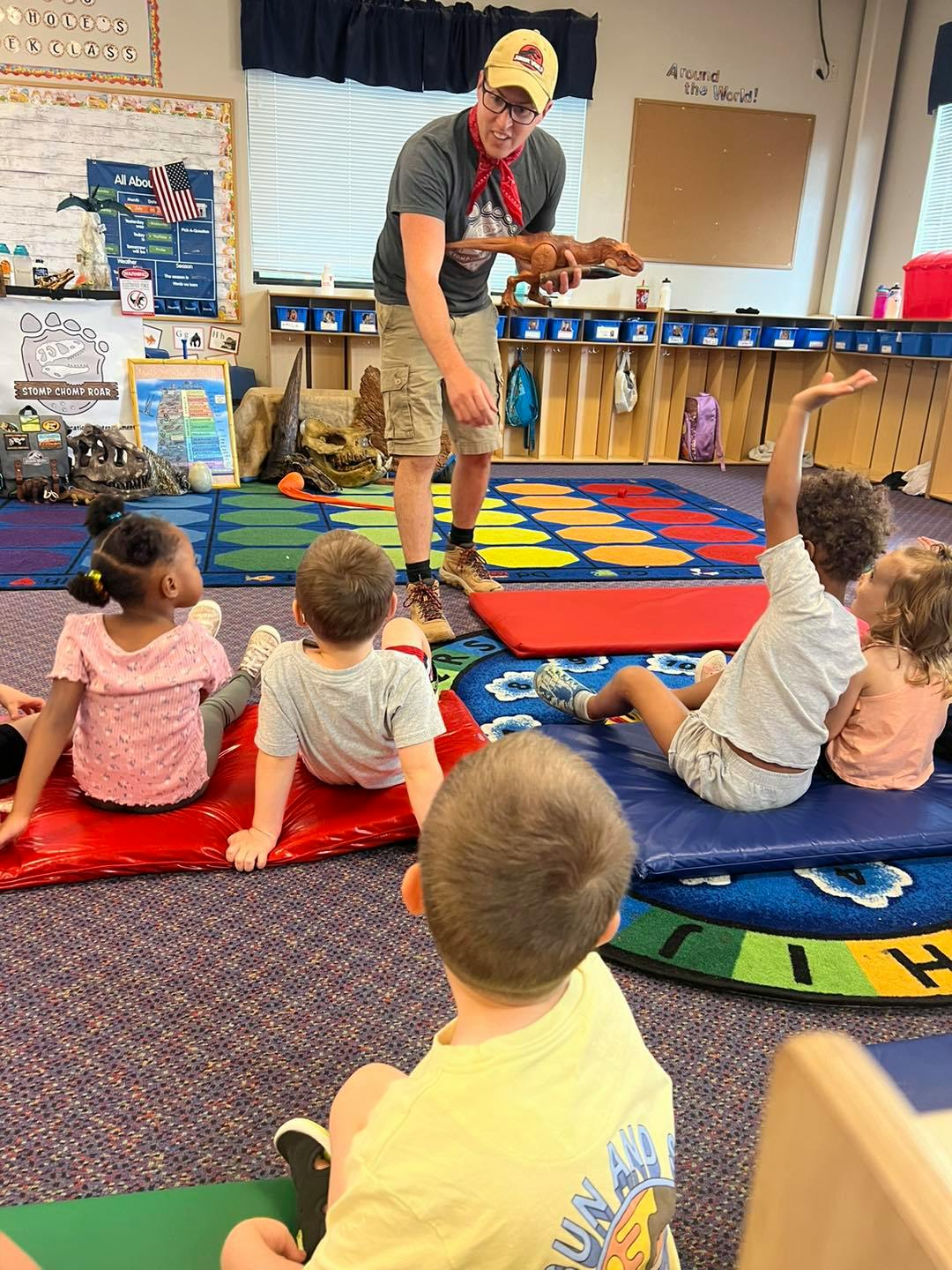 Adult holding a dinosaur toy for children sitting on mats in a classroom