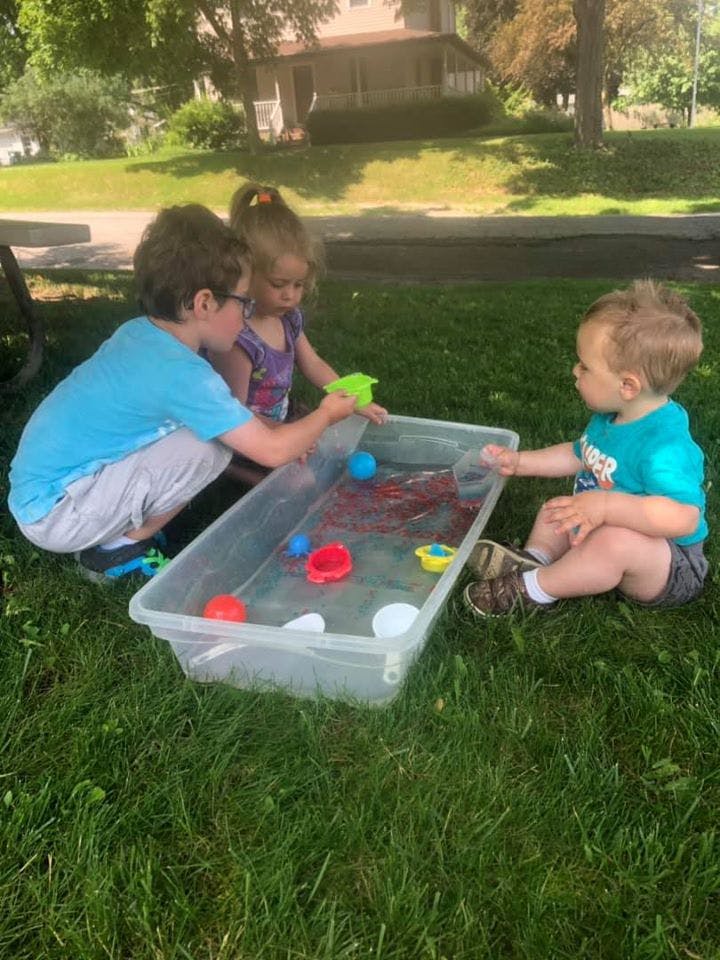Three children playing with water and toys in a plastic bin on grass