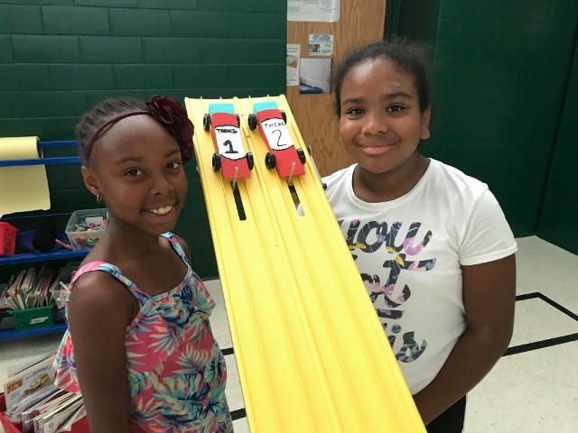 Two children standing next to a yellow ramp with two toy cars