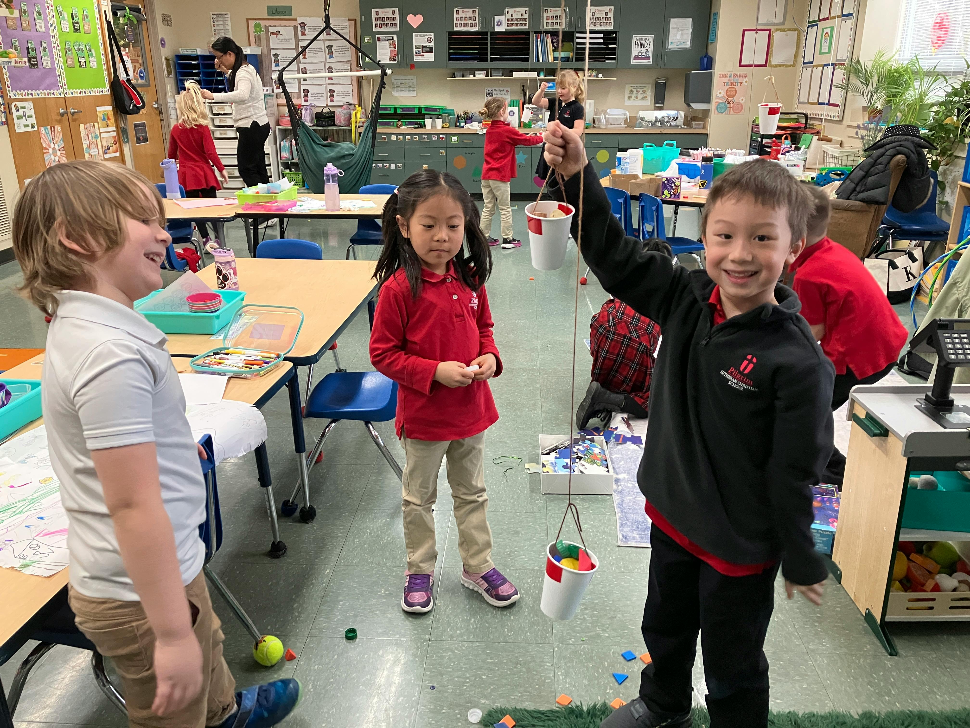 Children in a classroom, one child holds a cup attached to string, another stands nearby