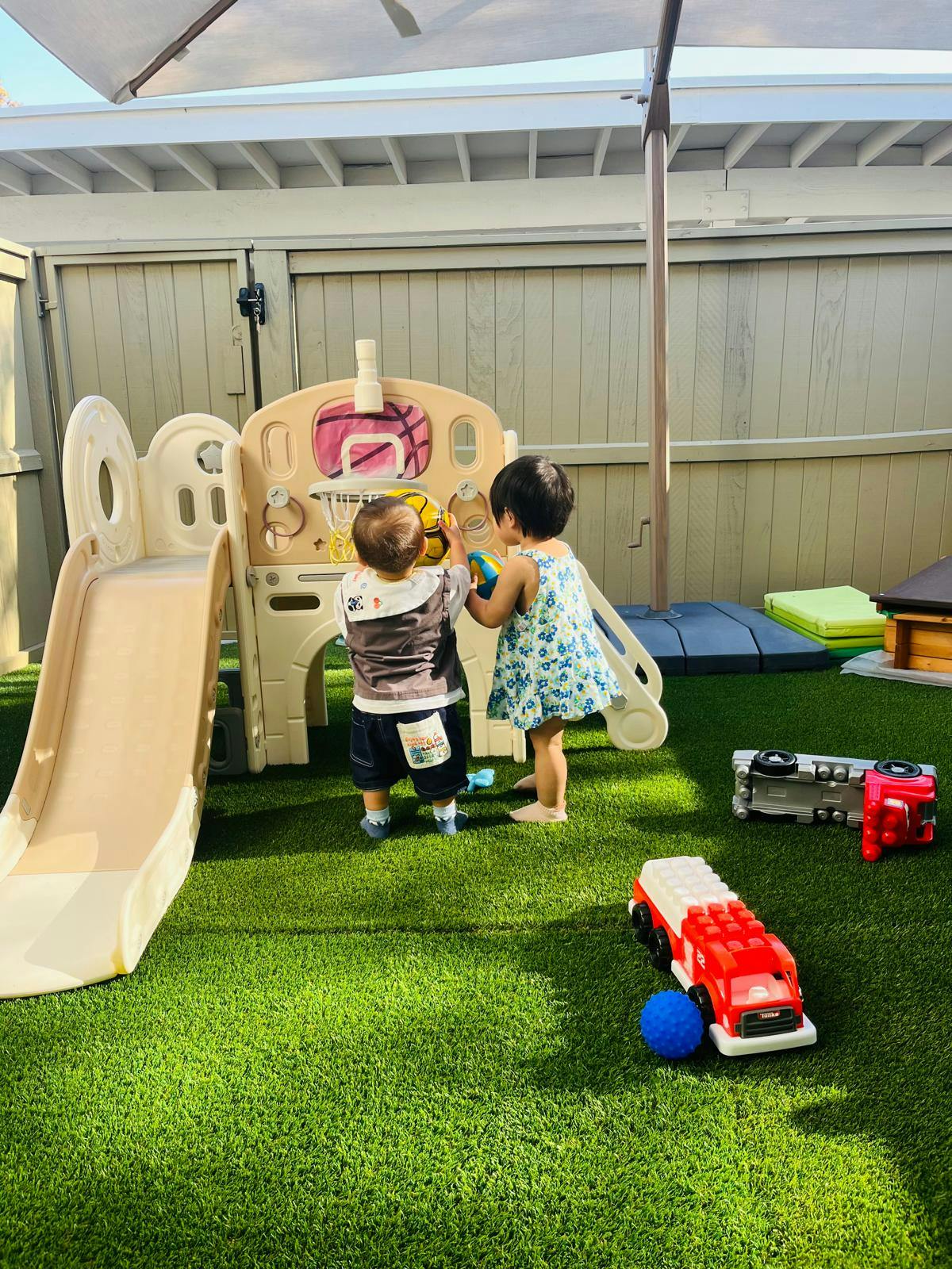 Two children playing with a toy basketball hoop and slide on artificial grass