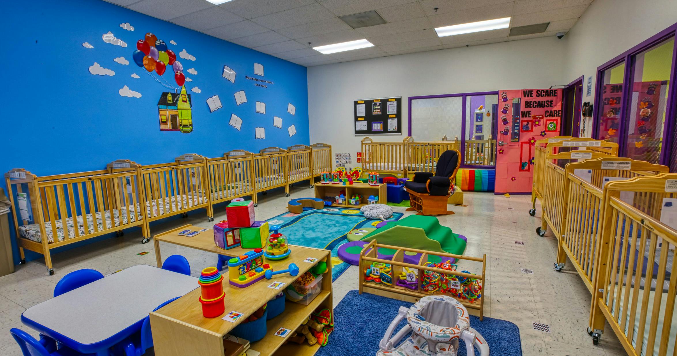 A colorful childcare classroom with cribs, play mats, and toys