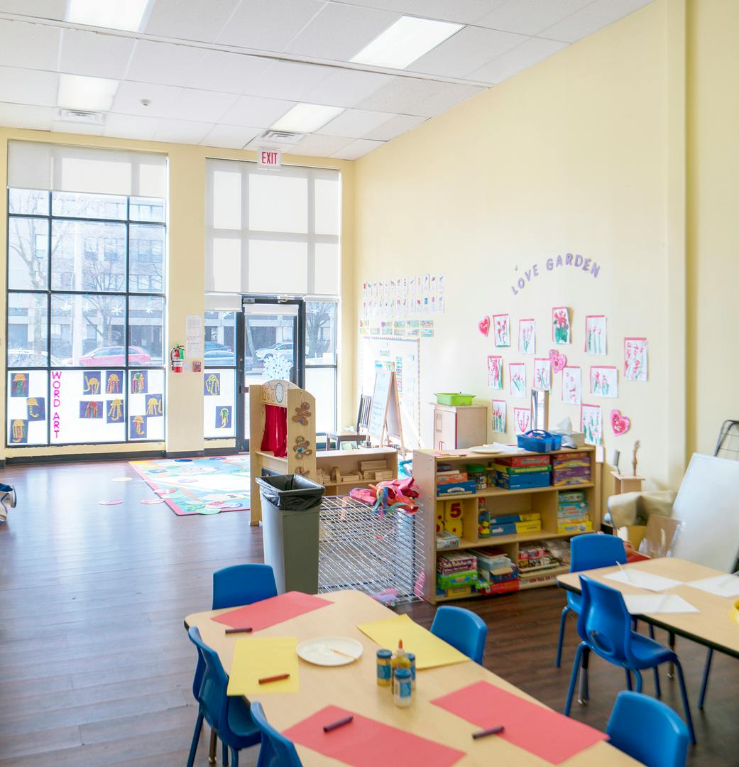 Classroom with tables and chairs, large windows, and children's artwork on wall