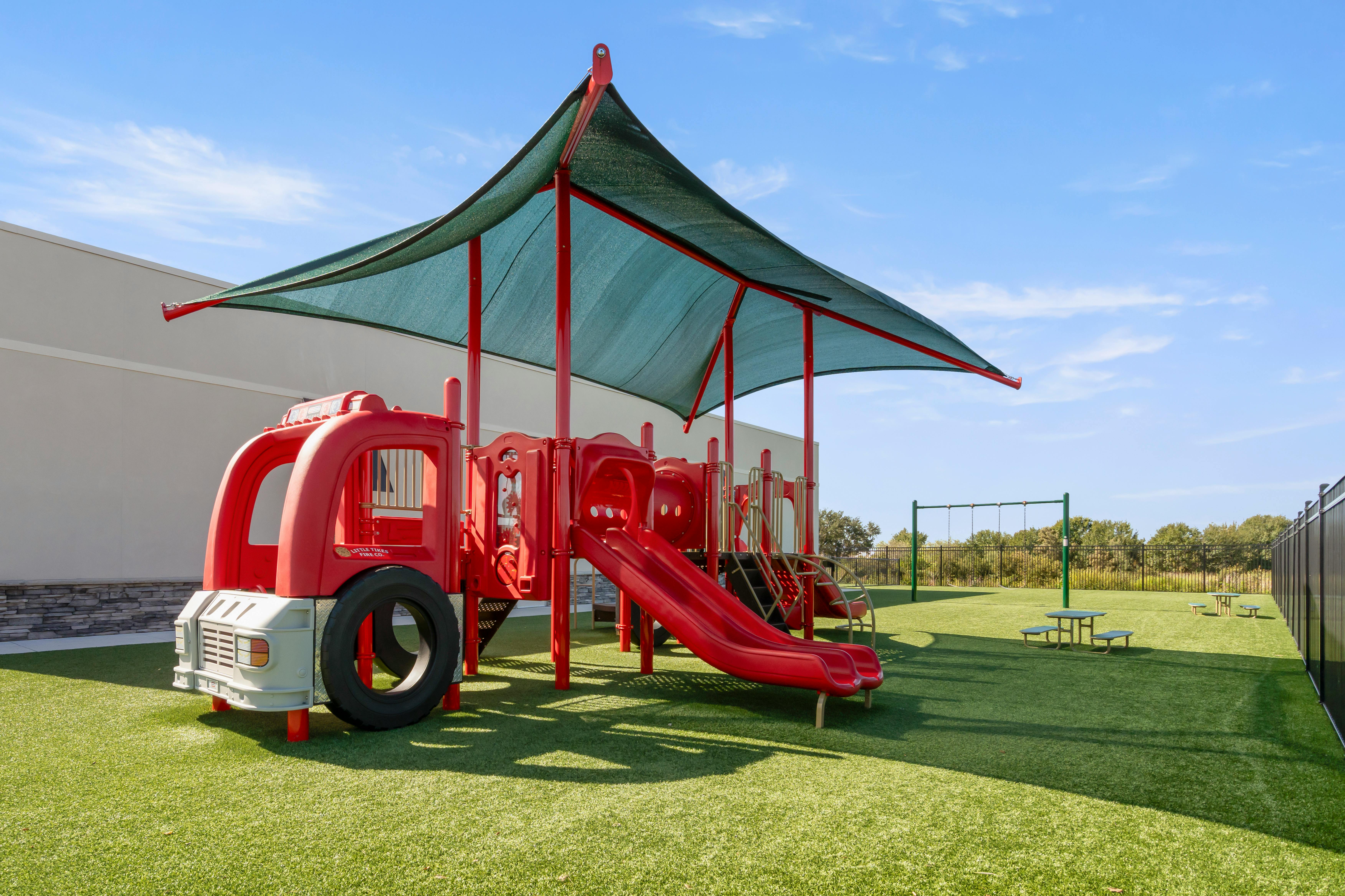 A red playground with slides and a fire truck shape under a green shade canopy