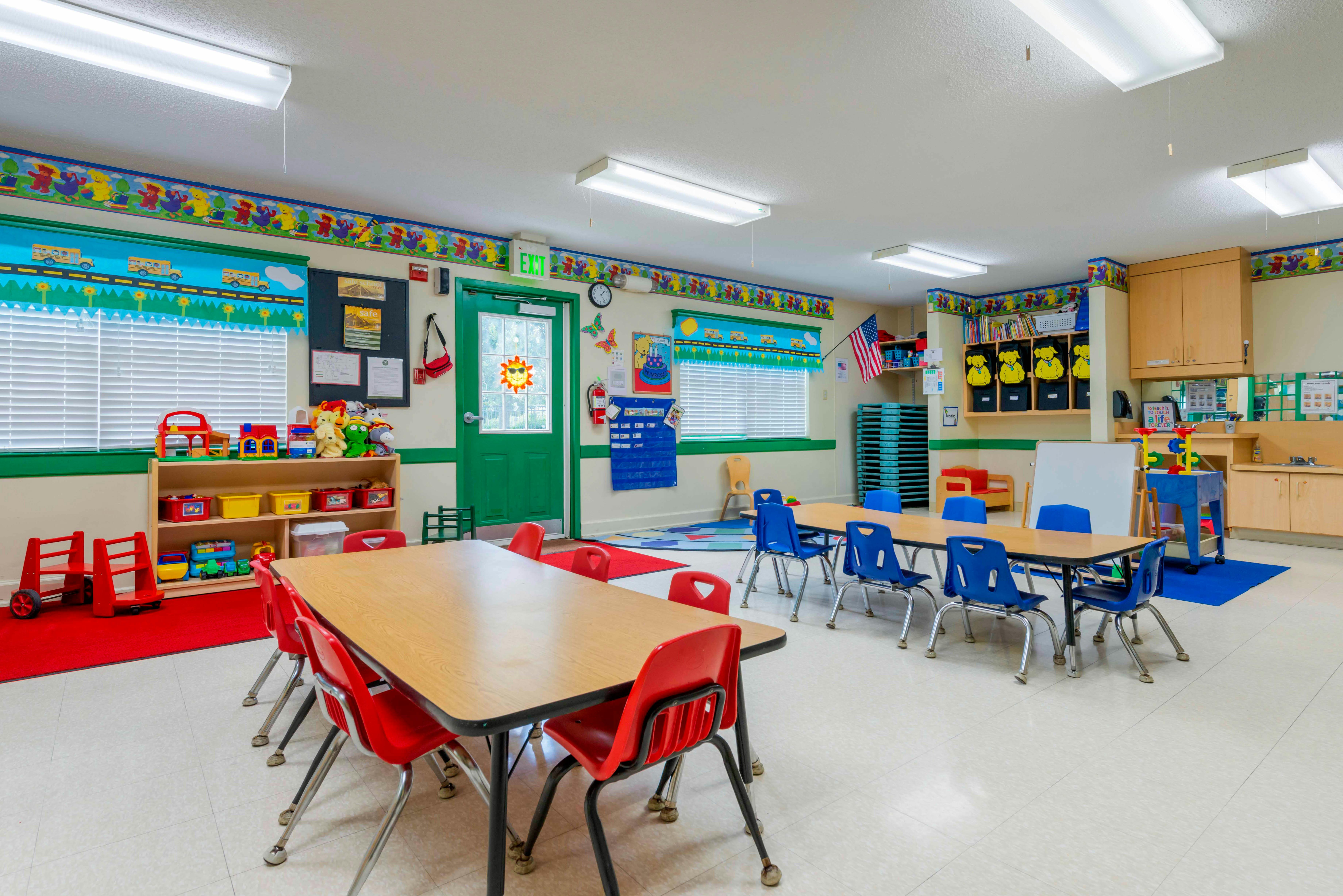 Empty classroom with small tables and chairs, toys on shelves, and an exit sign