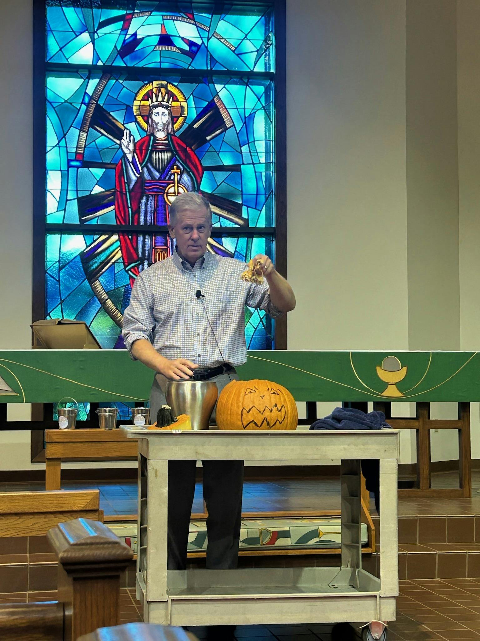 Person standing behind a table with a carved pumpkin in front of stained glass