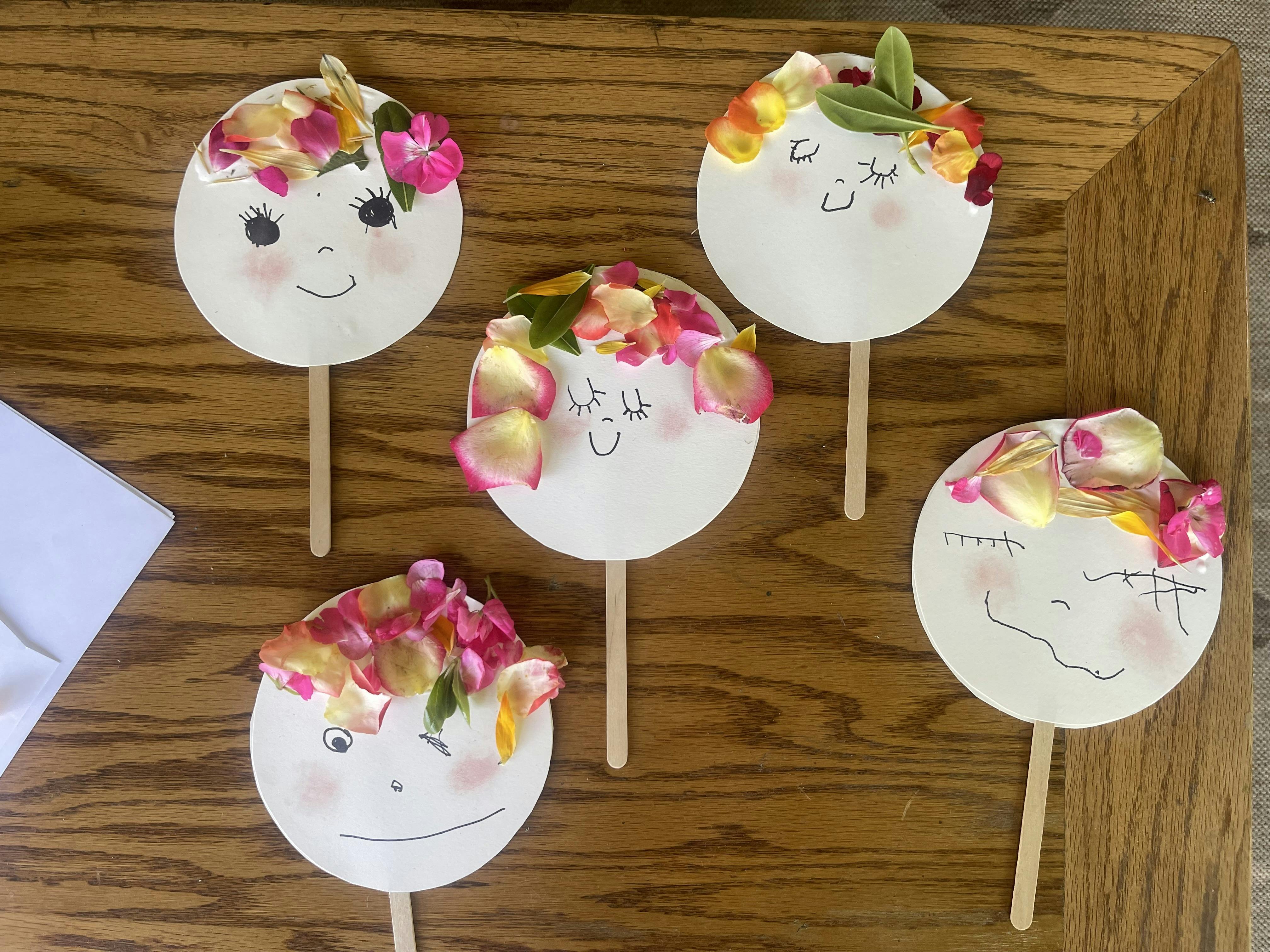 Five paper faces with drawn features and flower petal hair on sticks