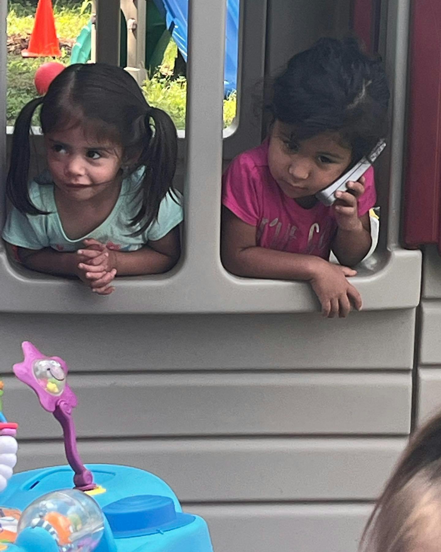 Two children looking out from a play structure, one holding a toy phone