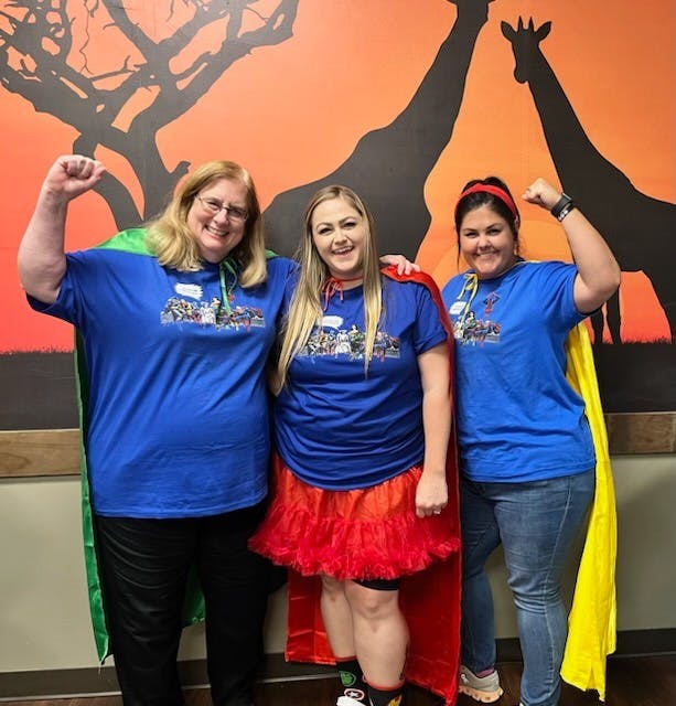 Three people in blue shirts and capes posing with fists raised