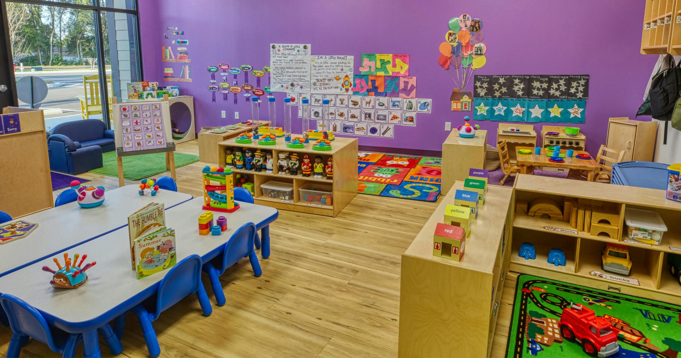 Colorful classroom with small tables and chairs, shelves with toys, and posters on purple walls