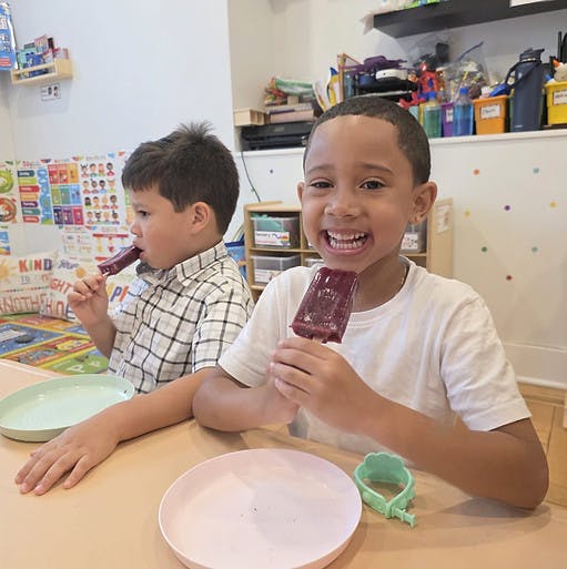 Two children eating popsicles at a table in a classroom