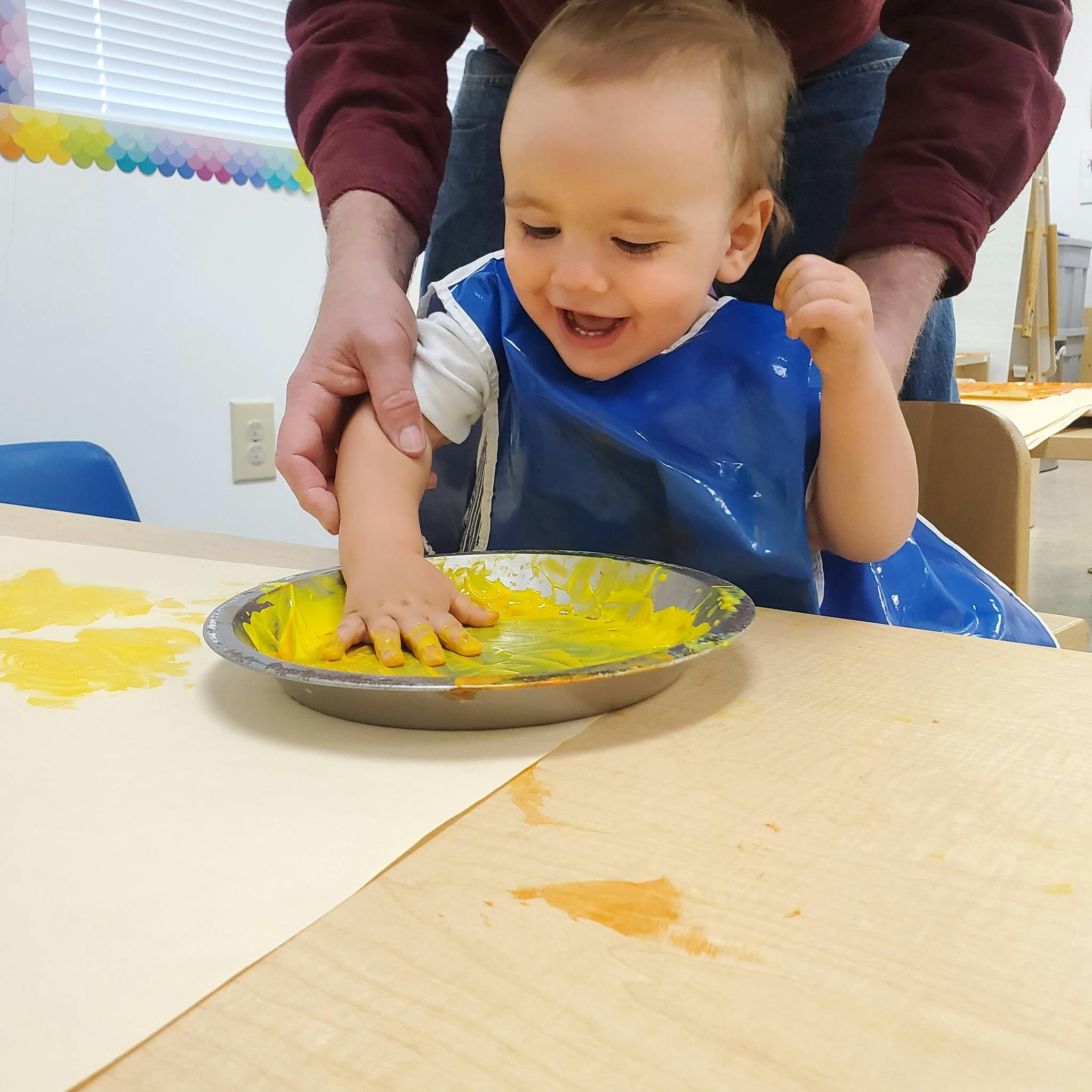 Child in a blue bib with a person's hands helping them finger paint with yellow paint