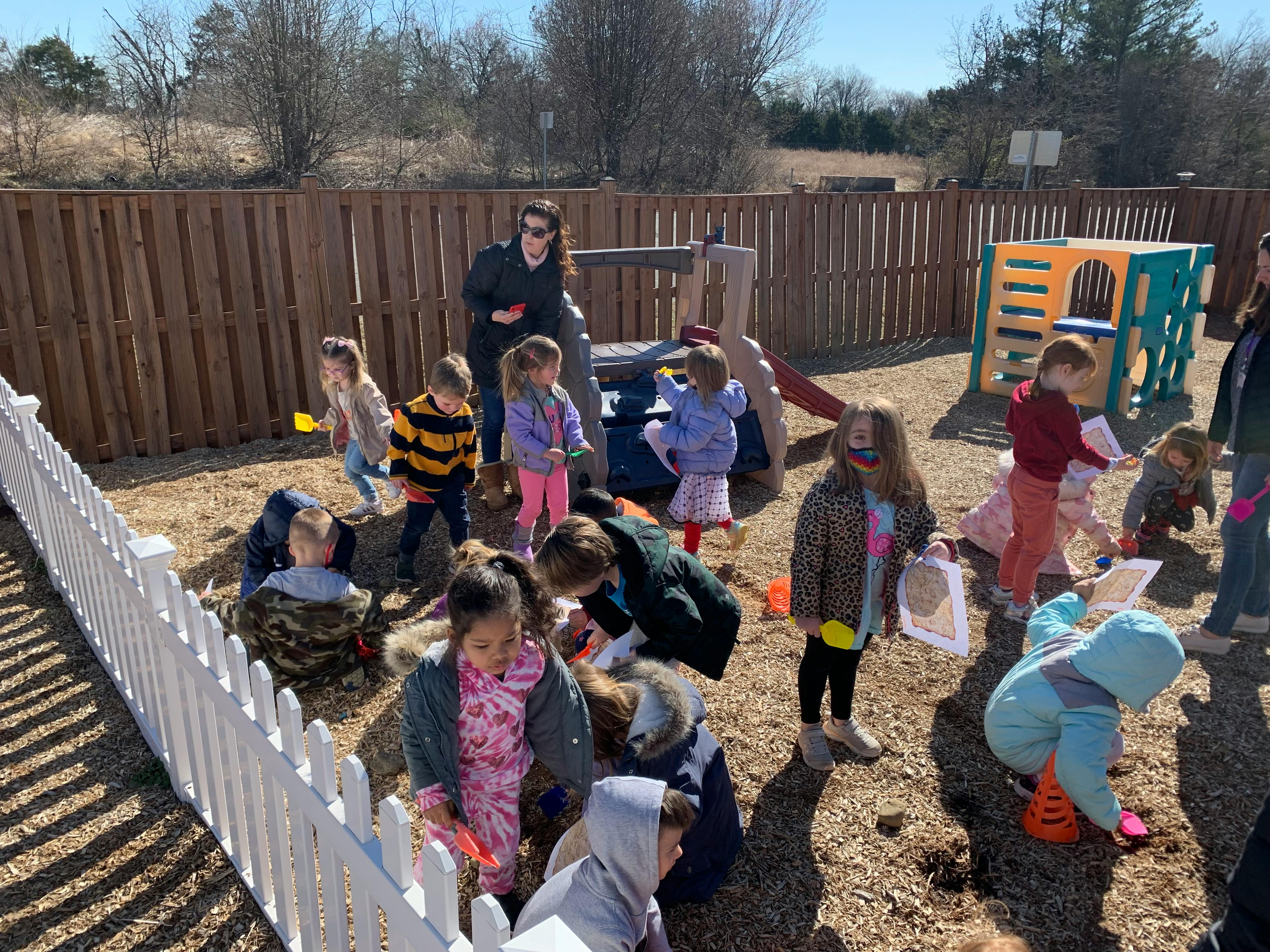 Adults and children playing in a dirt area with a fence and playground equipment