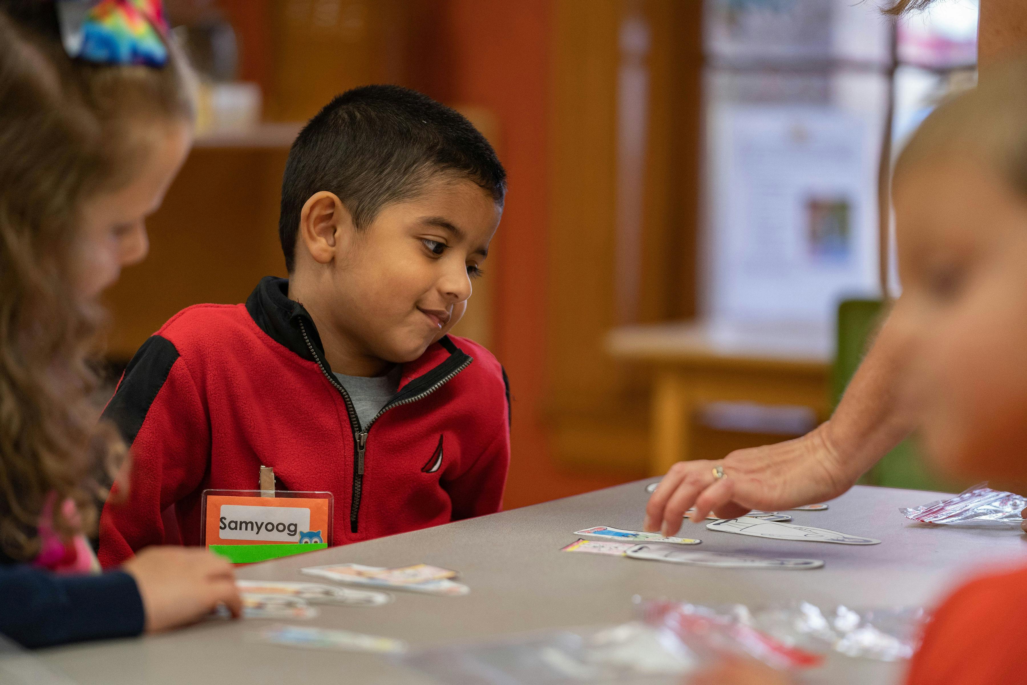 Child in red jacket looking at items on a table with other children