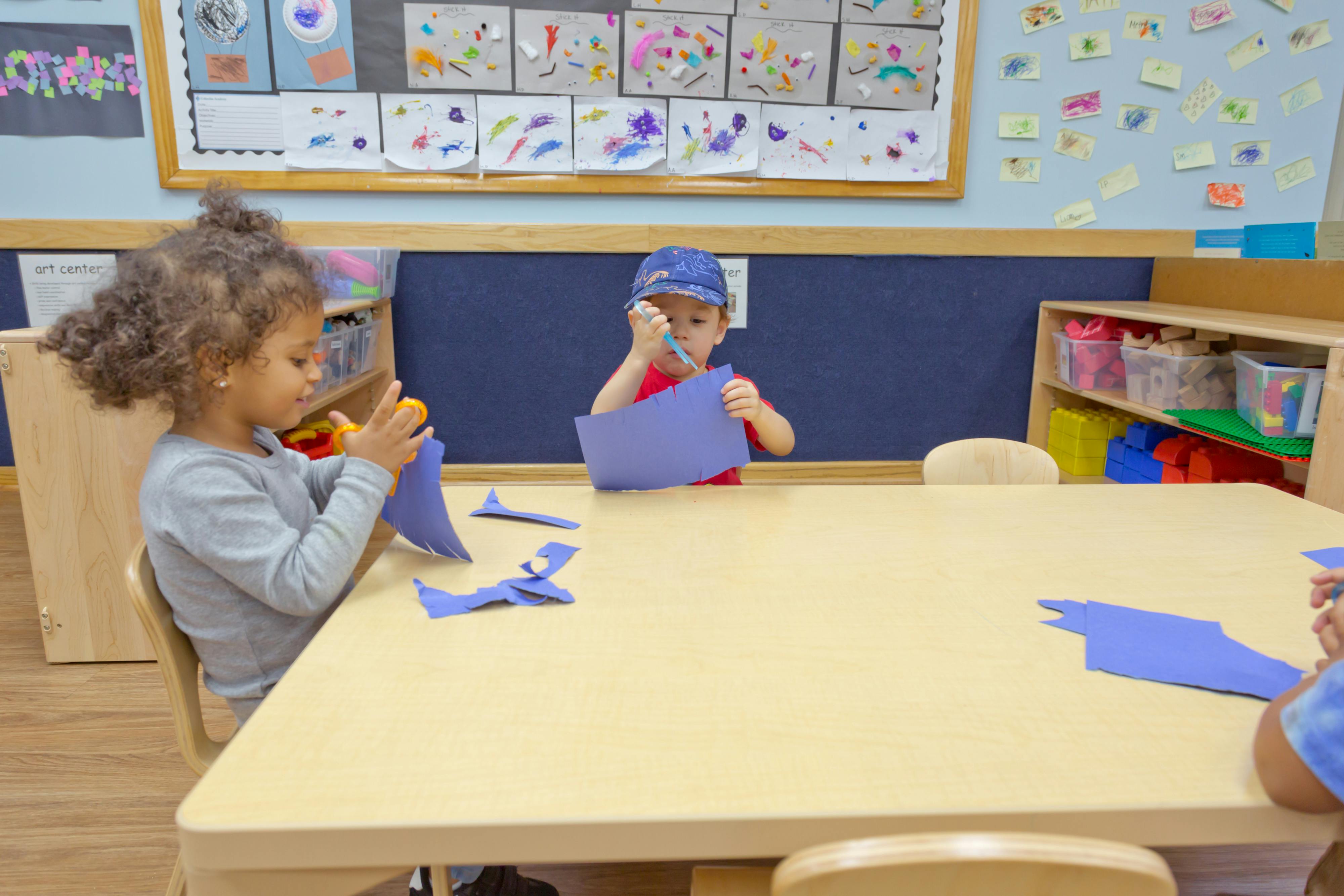 Two children cutting blue paper at a table in a classroom