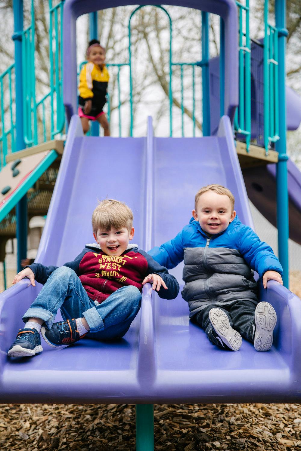 Two children sitting on a purple slide, another child standing at the top