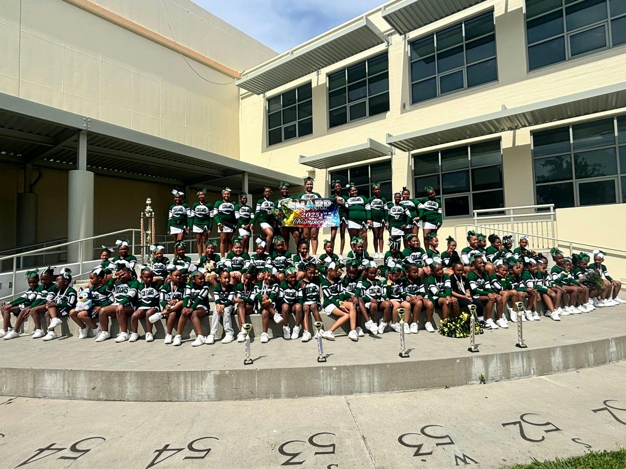Group of cheerleaders wearing green and white uniforms with trophies and a banner