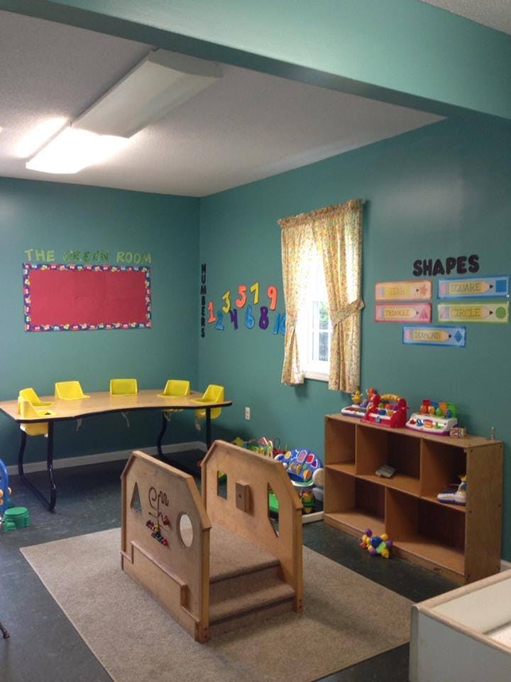 Classroom with a long table and yellow chairs, play structure, and shelves with toys