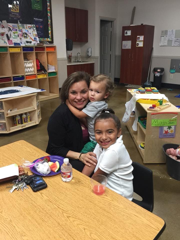 Person holding a child, with another child sitting at a table in a classroom