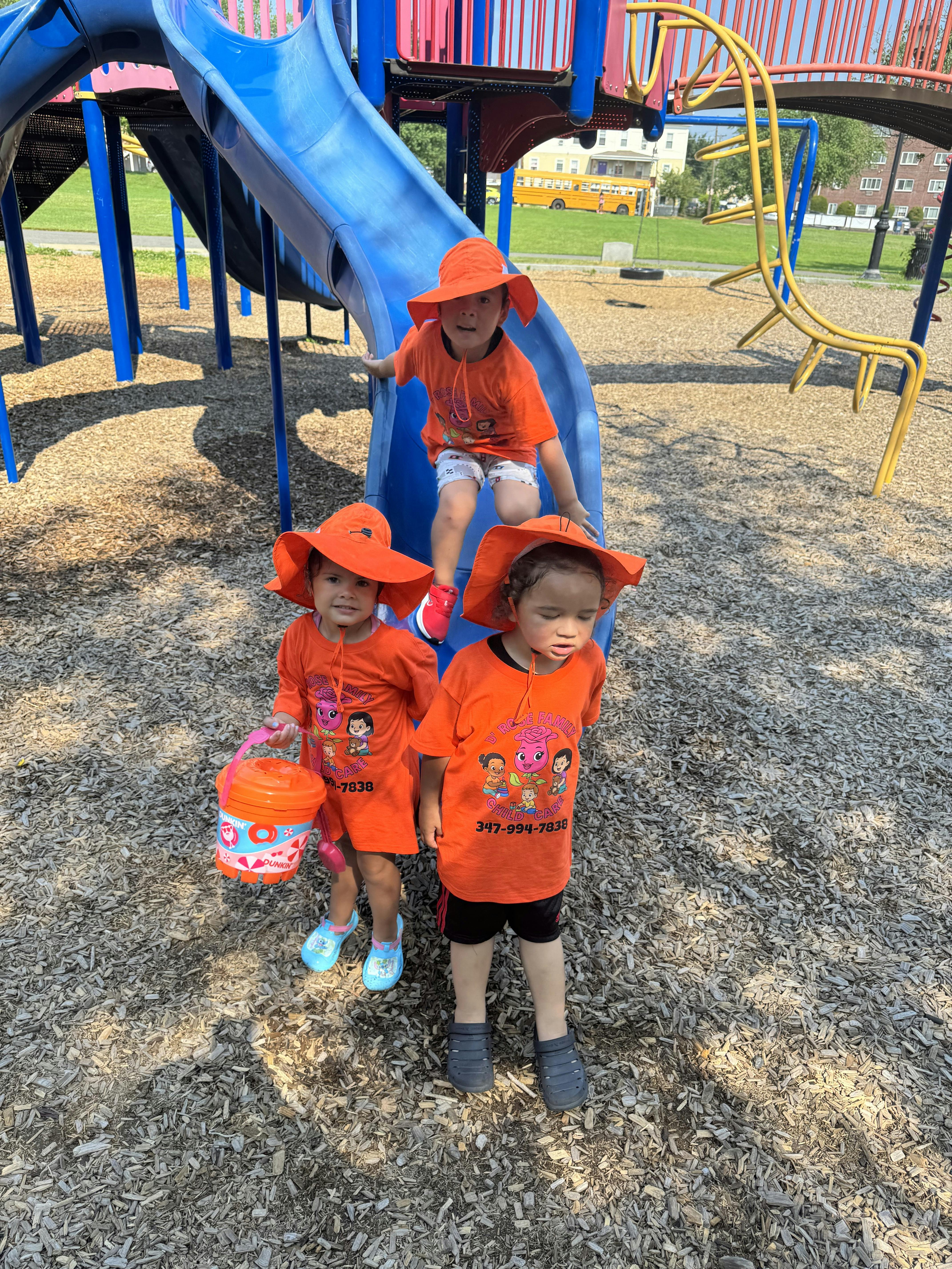 Three children in orange shirts and hats on a playground slide