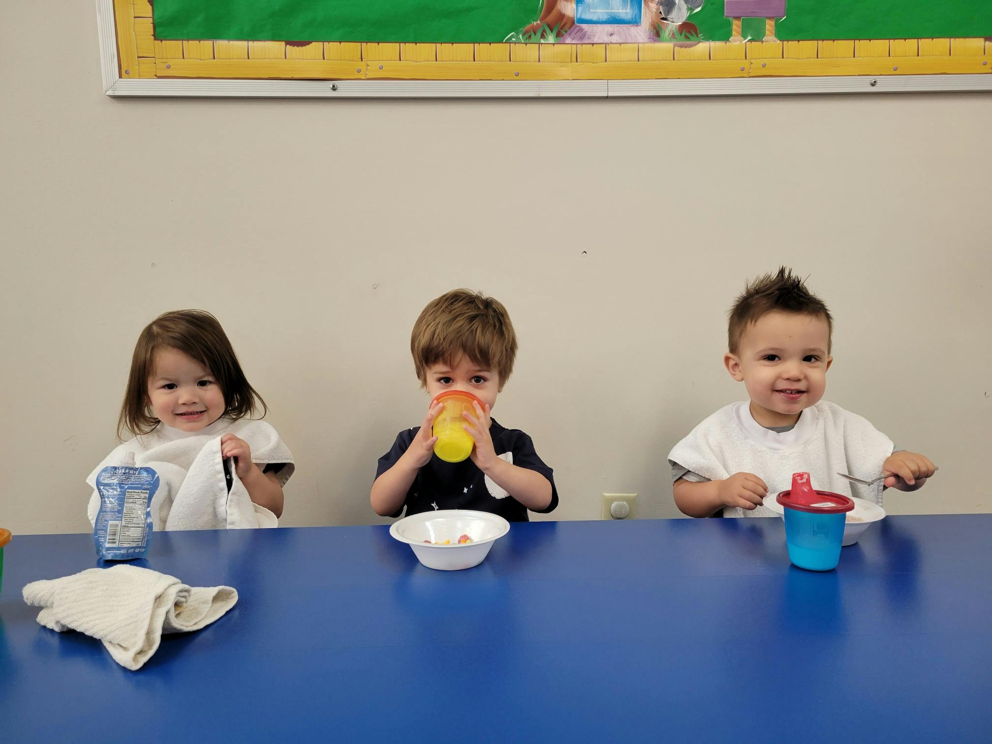 Three children sitting at a table, one drinking from a cup