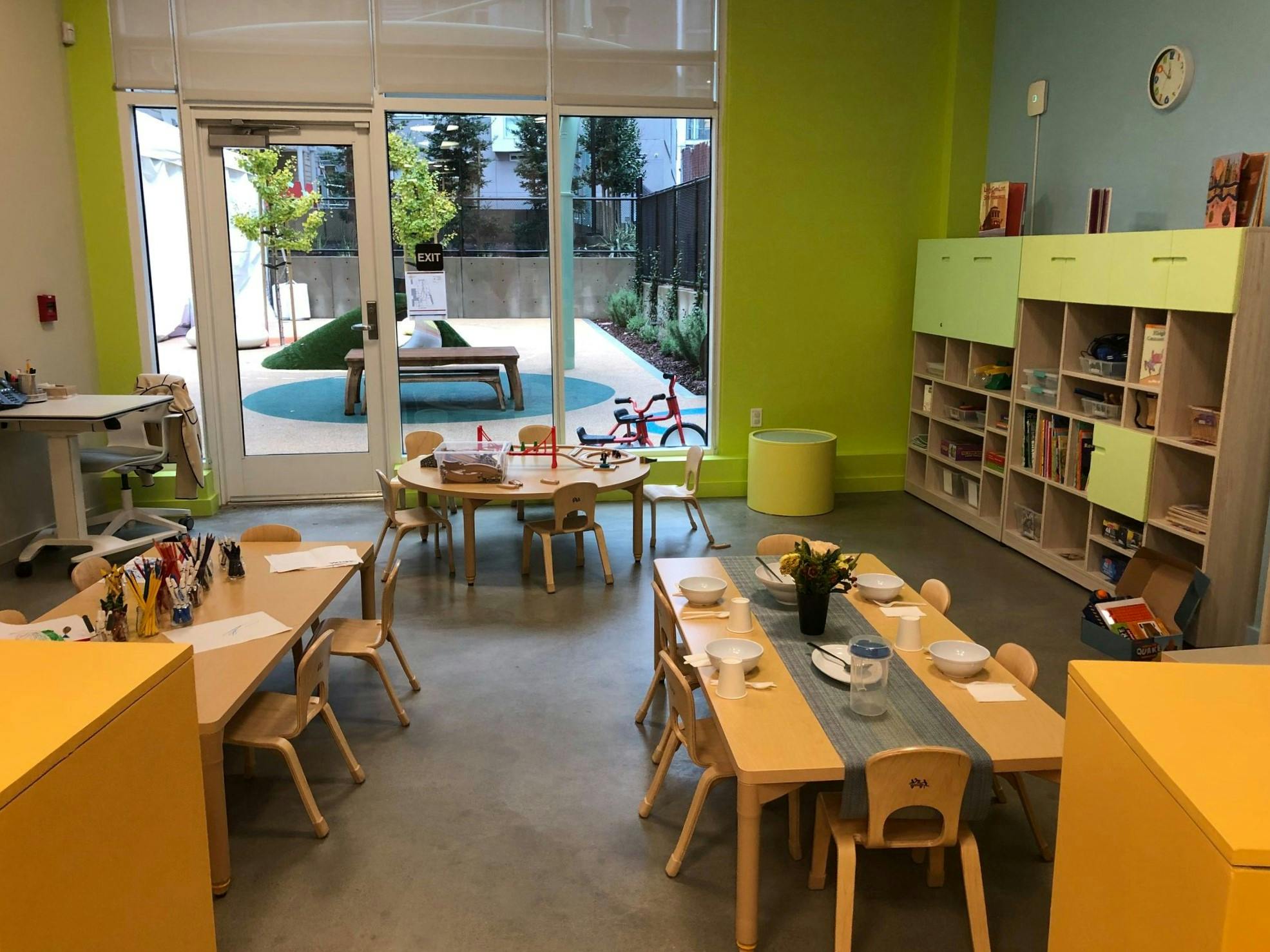 Classroom with small tables and chairs, bookshelves, and a view to an outdoor play area
