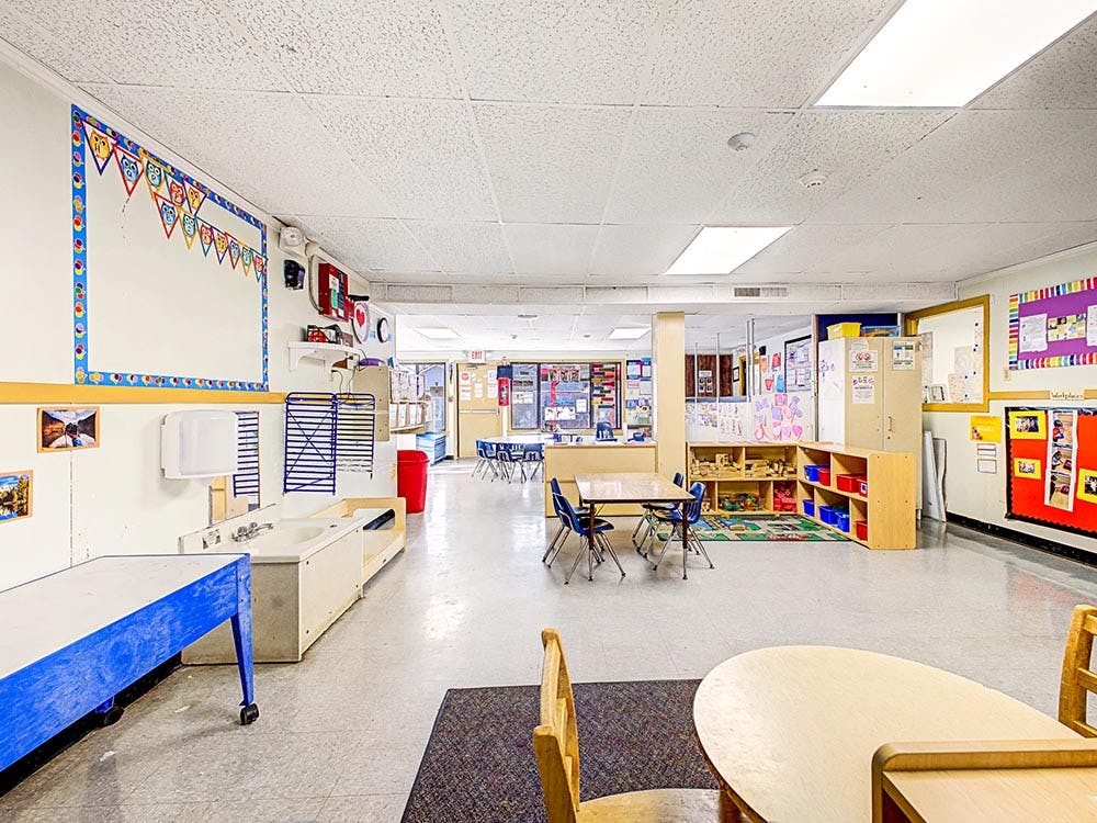 Empty classroom with small tables and chairs, shelves with toys, and decorated walls