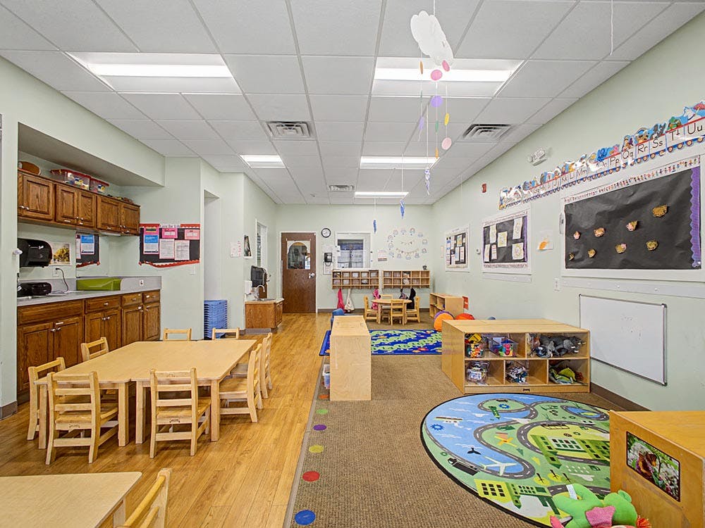 Empty classroom with small wooden tables and chairs, play mats, and cabinets