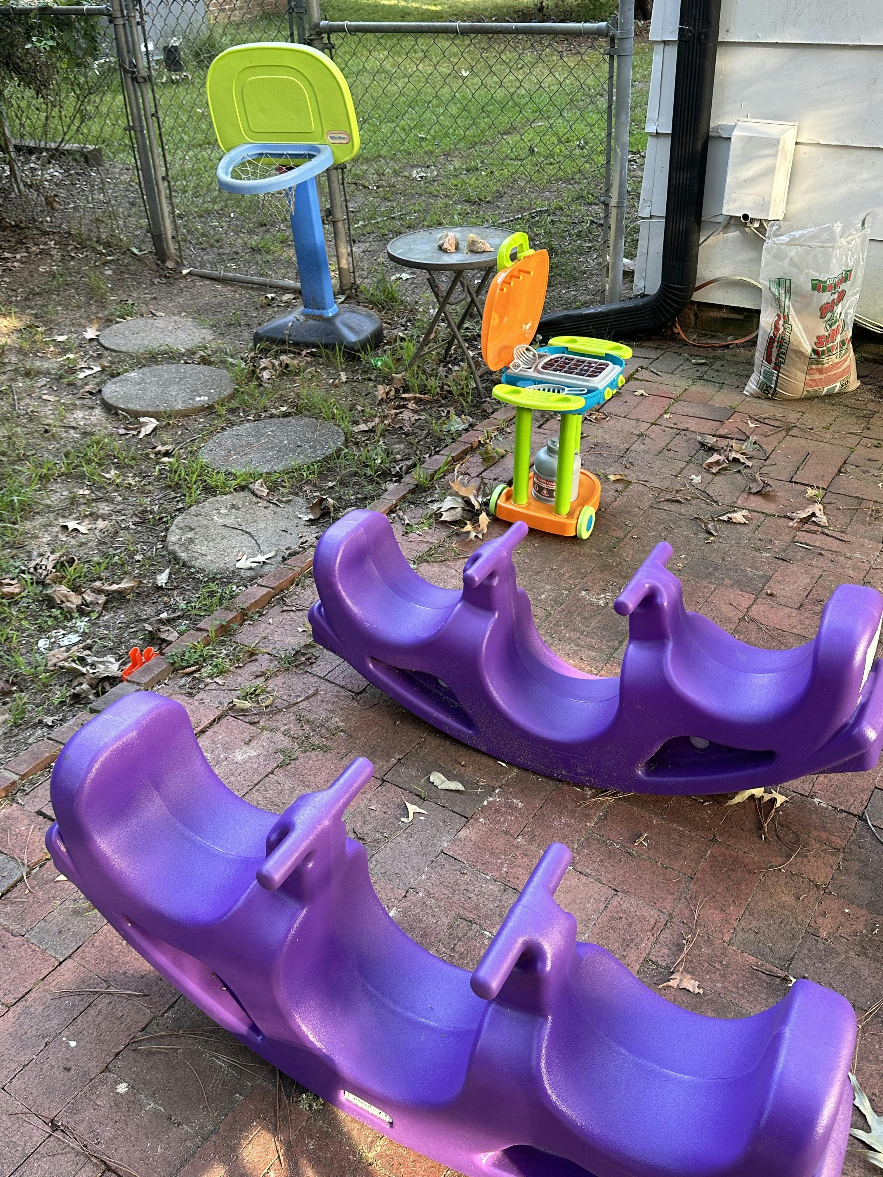 Two purple plastic seesaws on a brick patio with other toys in the background