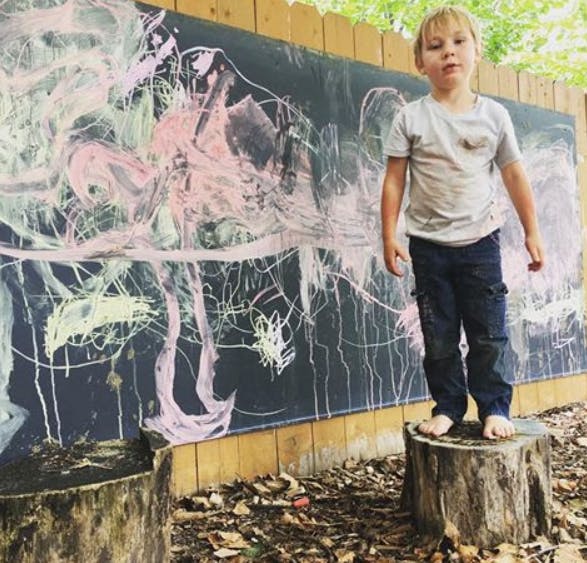 Child standing barefoot on a tree stump in front of a chalk-covered wall