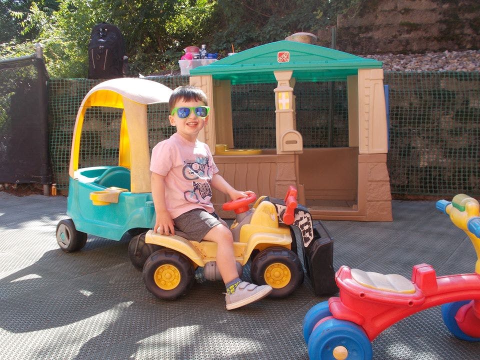 Child in sunglasses sitting on a toy tractor next to a toy car