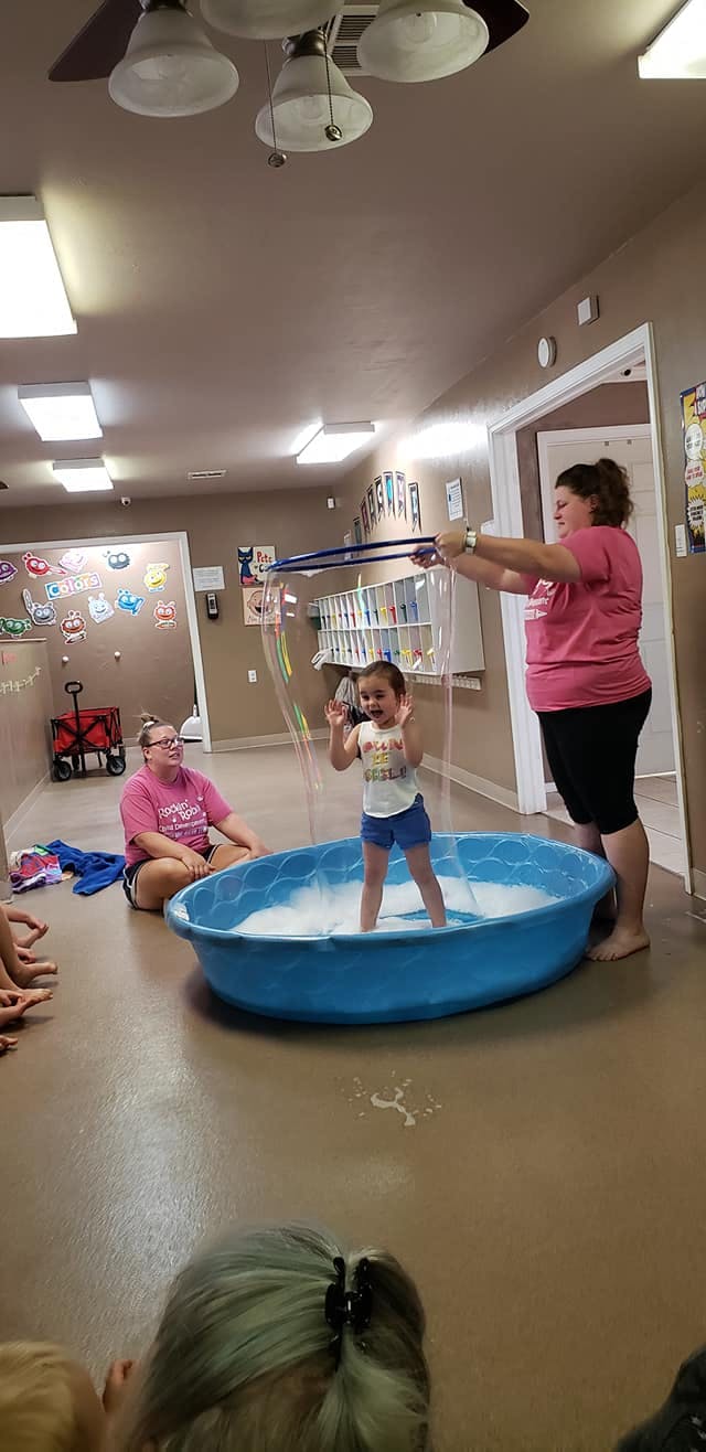 Person making a large bubble around a child standing in a blue tub
