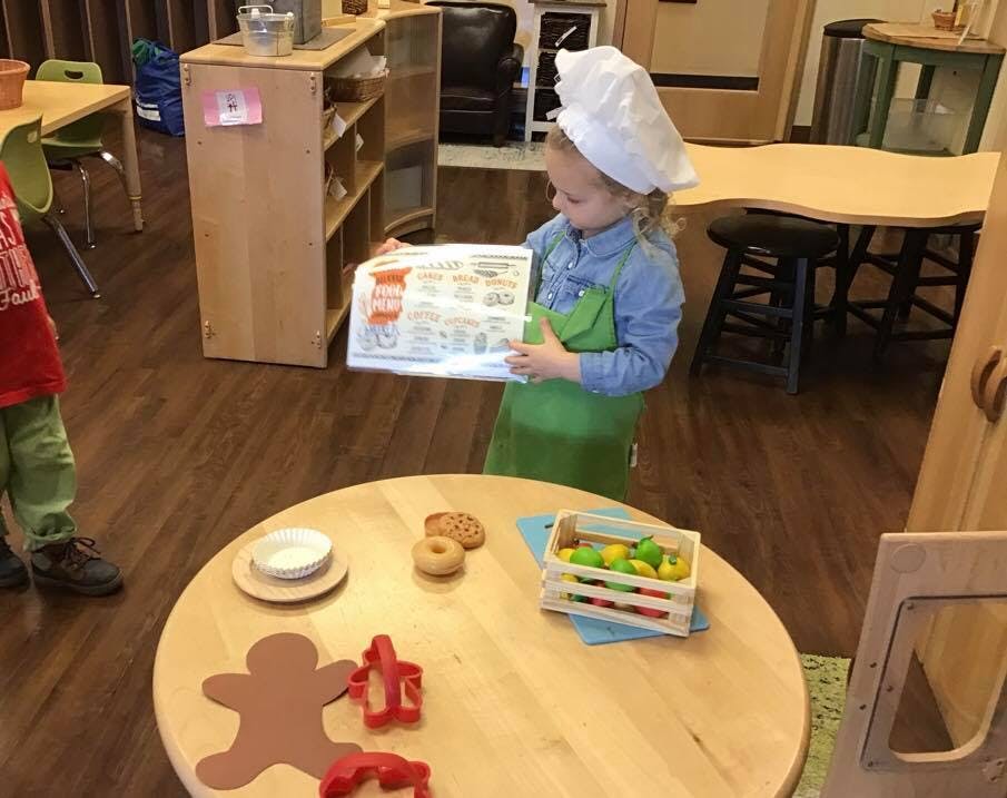 Child in chef hat and apron holding a book over a table with play food