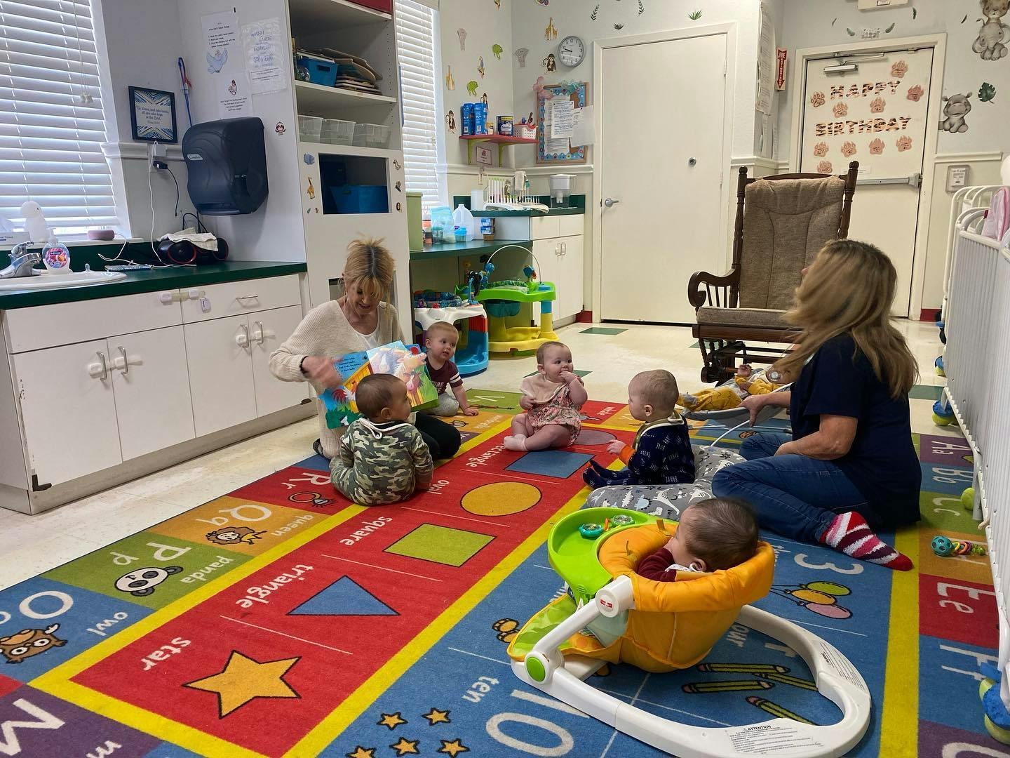 Two people interacting with multiple babies on a colorful rug in a room