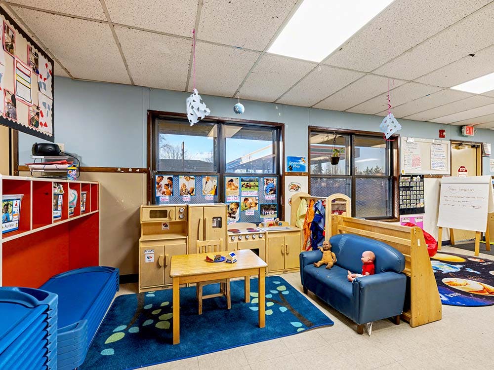 A classroom with a play kitchen set, small table, couch with dolls, and blue mats
