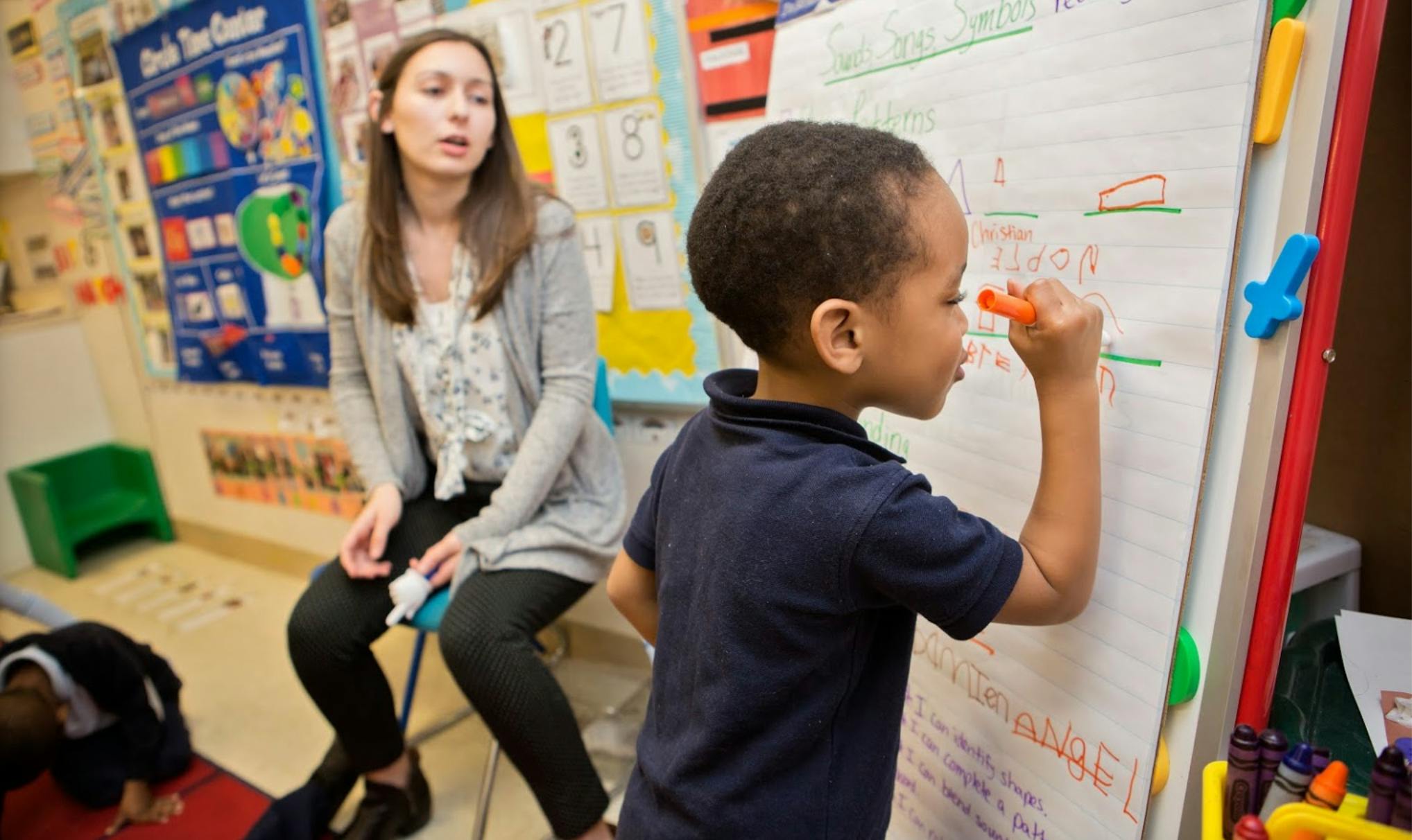 Child drawing on a whiteboard with a person sitting nearby