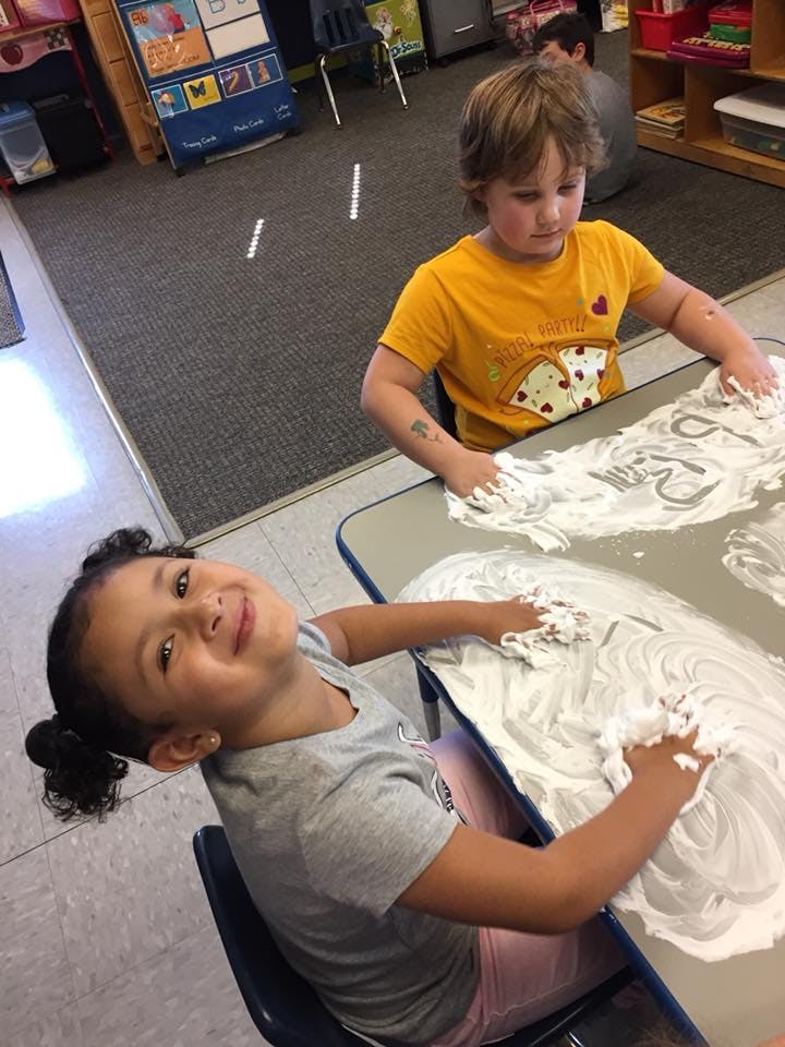 Two children playing with shaving cream on a table in a classroom