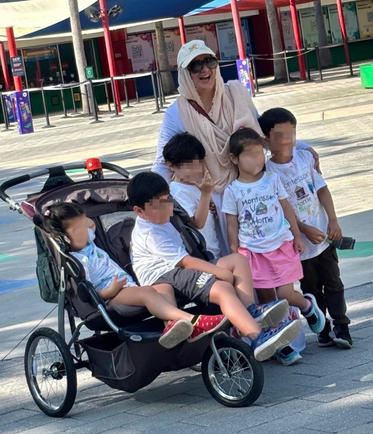 Person in a head covering and sunglasses with five children, two in a stroller