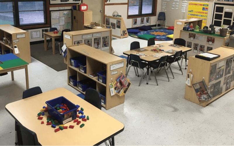 An empty classroom with several small tables and chairs, shelves, and a rug